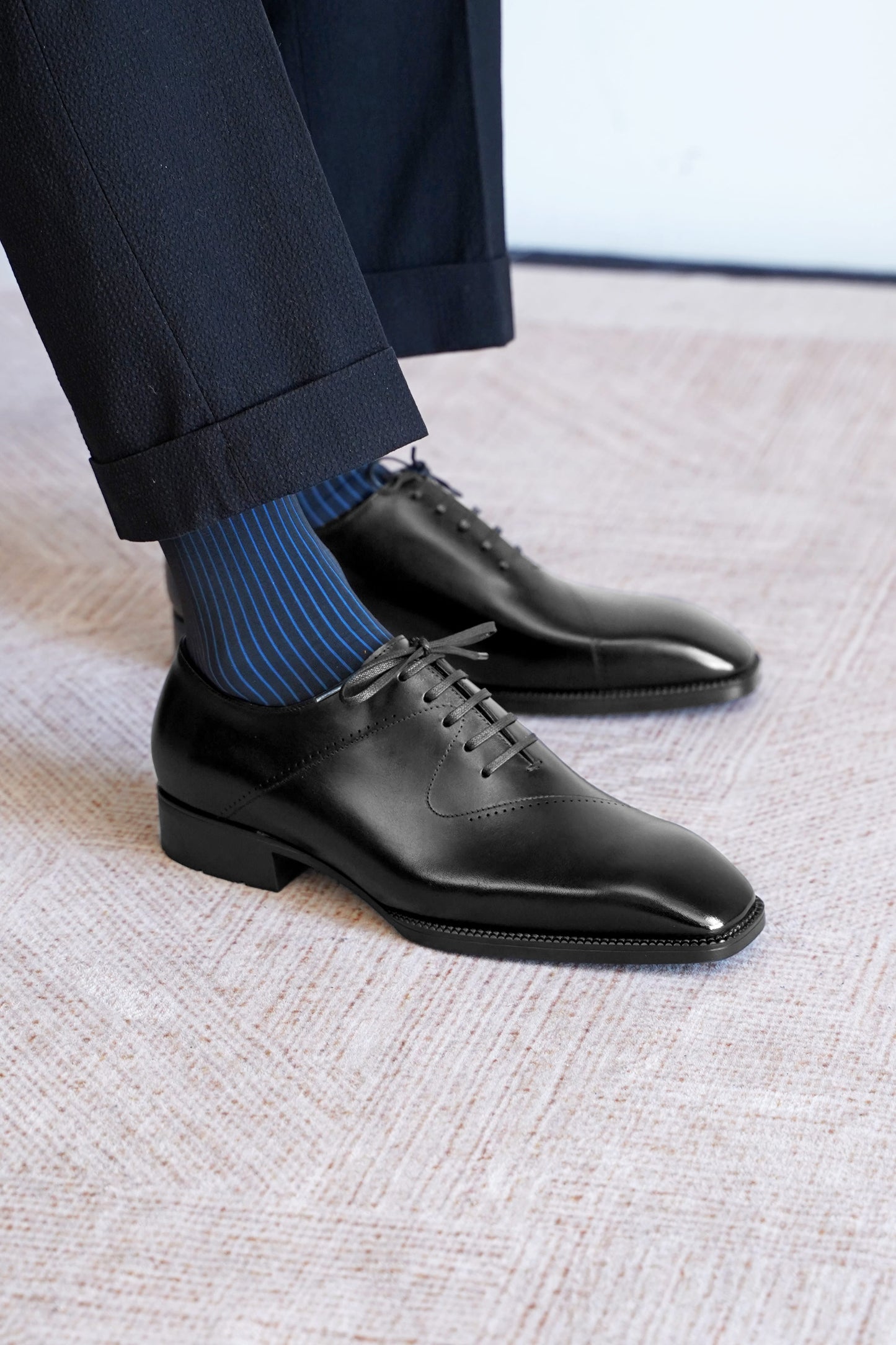 A close-up of a person's feet wearing black leather oxford dress shoes with blue and navy striped socks, paired with dark trousers, standing on a textured light beige carpet.