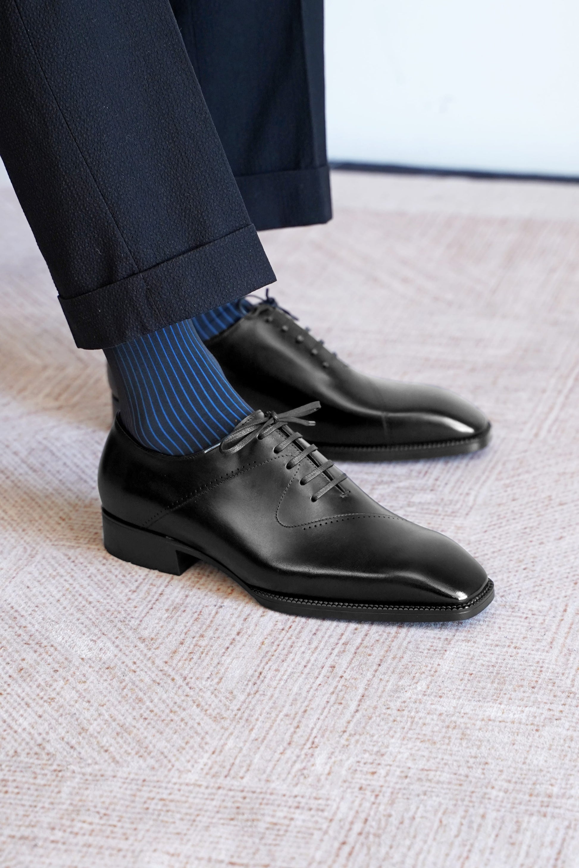 A close-up of a person's feet wearing black leather oxford dress shoes with blue and navy striped socks, paired with dark trousers, standing on a textured light beige carpet.