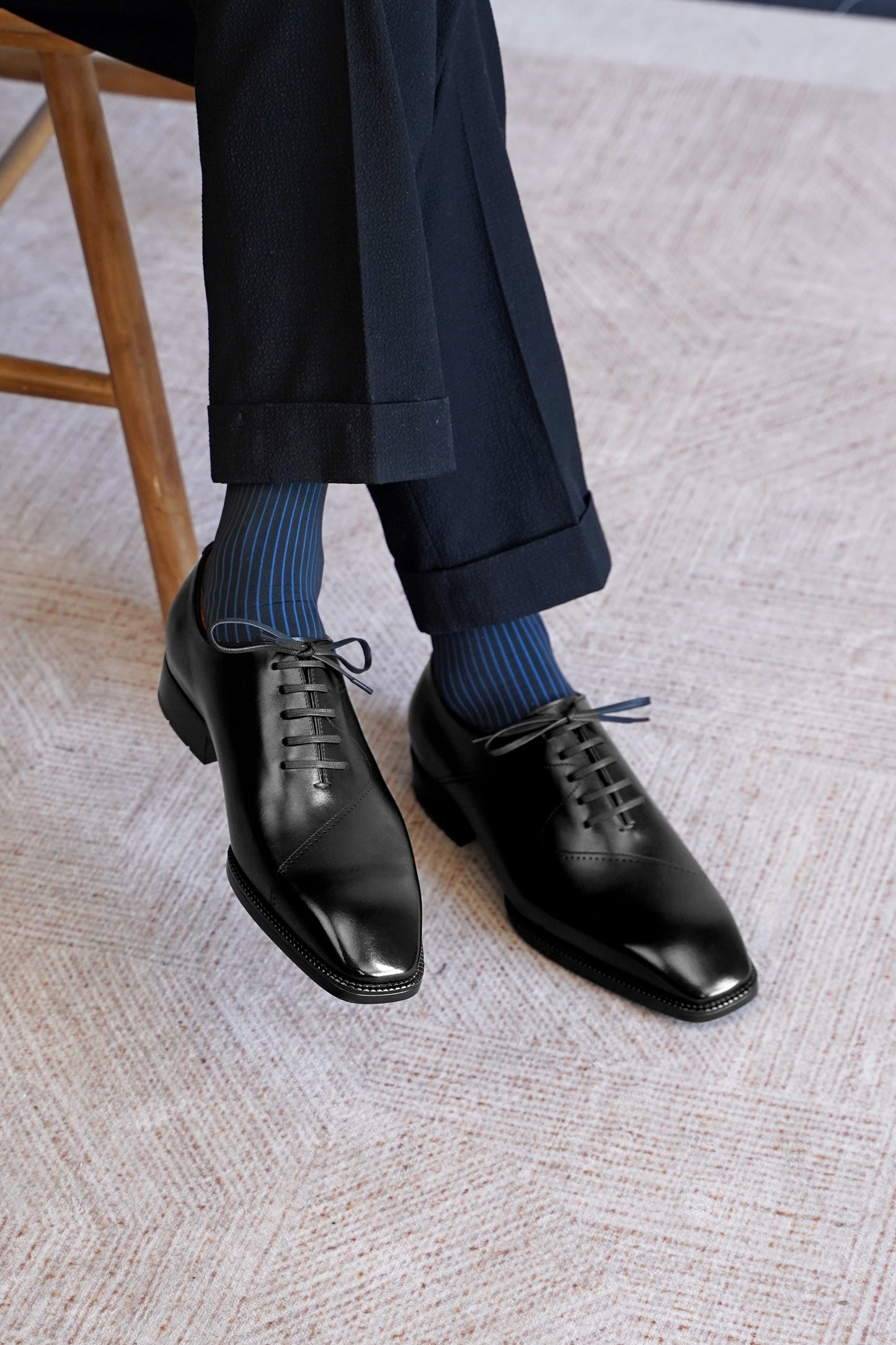 A close-up of a person's legs wearing black leather oxford dress shoes, dark navy trousers, and blue striped socks, sitting on a textured beige carpet next to a wooden chair.
