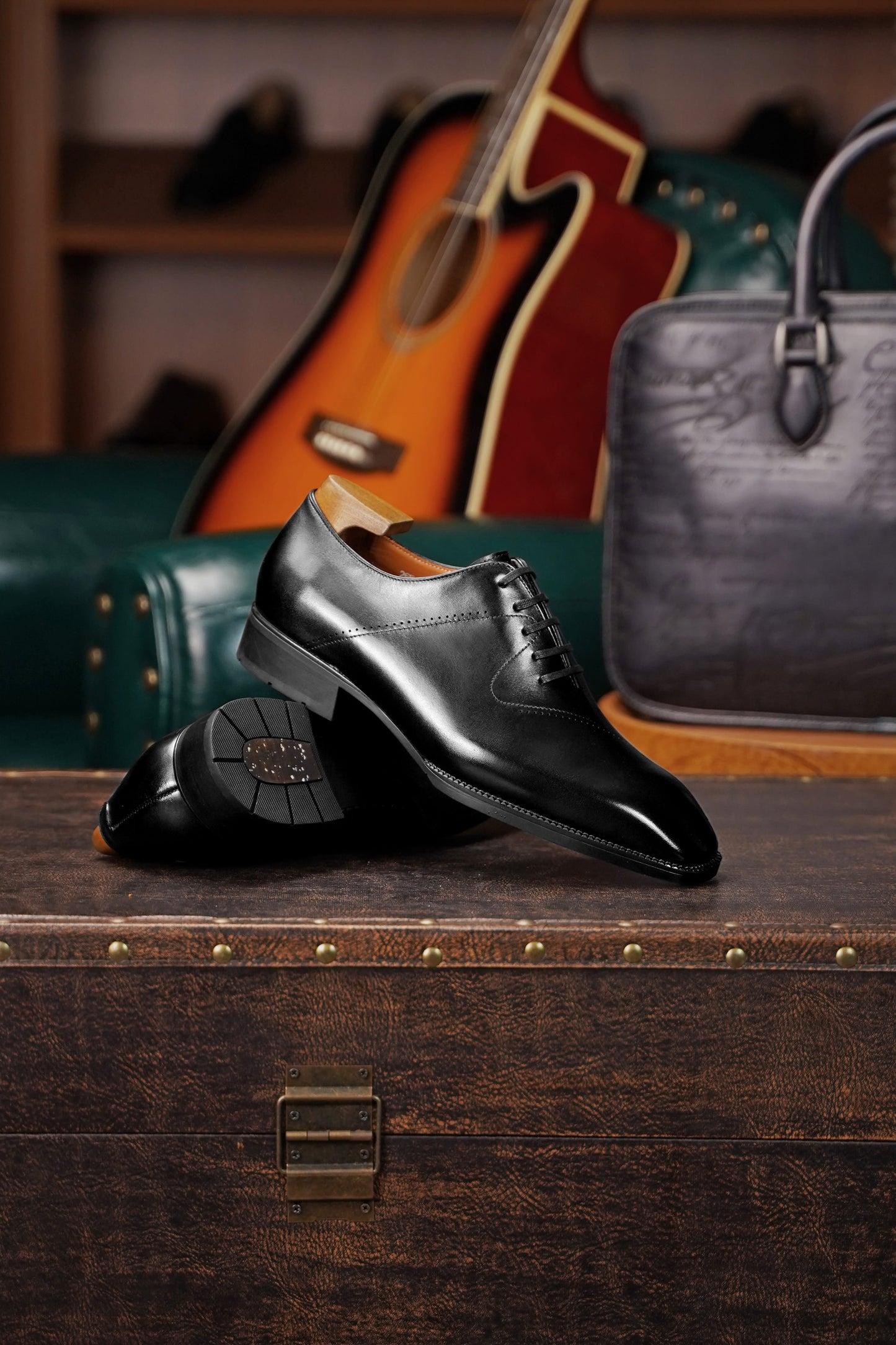 A pair of polished black leather oxford dress shoes displayed on a rustic wooden trunk, with a green leather sofa, an acoustic guitar, and a dark gray textured handbag in the softly blurred background.