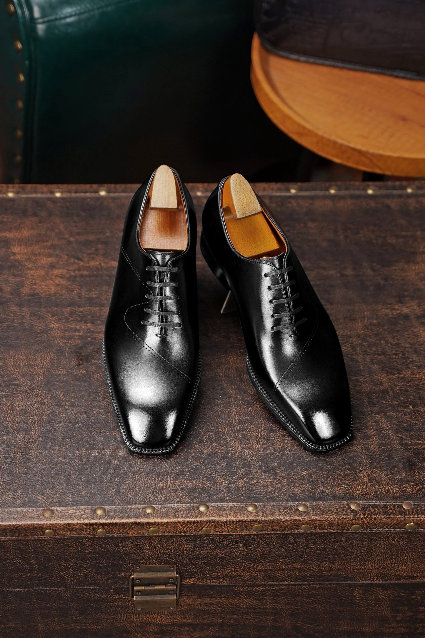 A pair of black leather lace-up dress shoes with a polished finish, displayed on a dark wooden trunk.