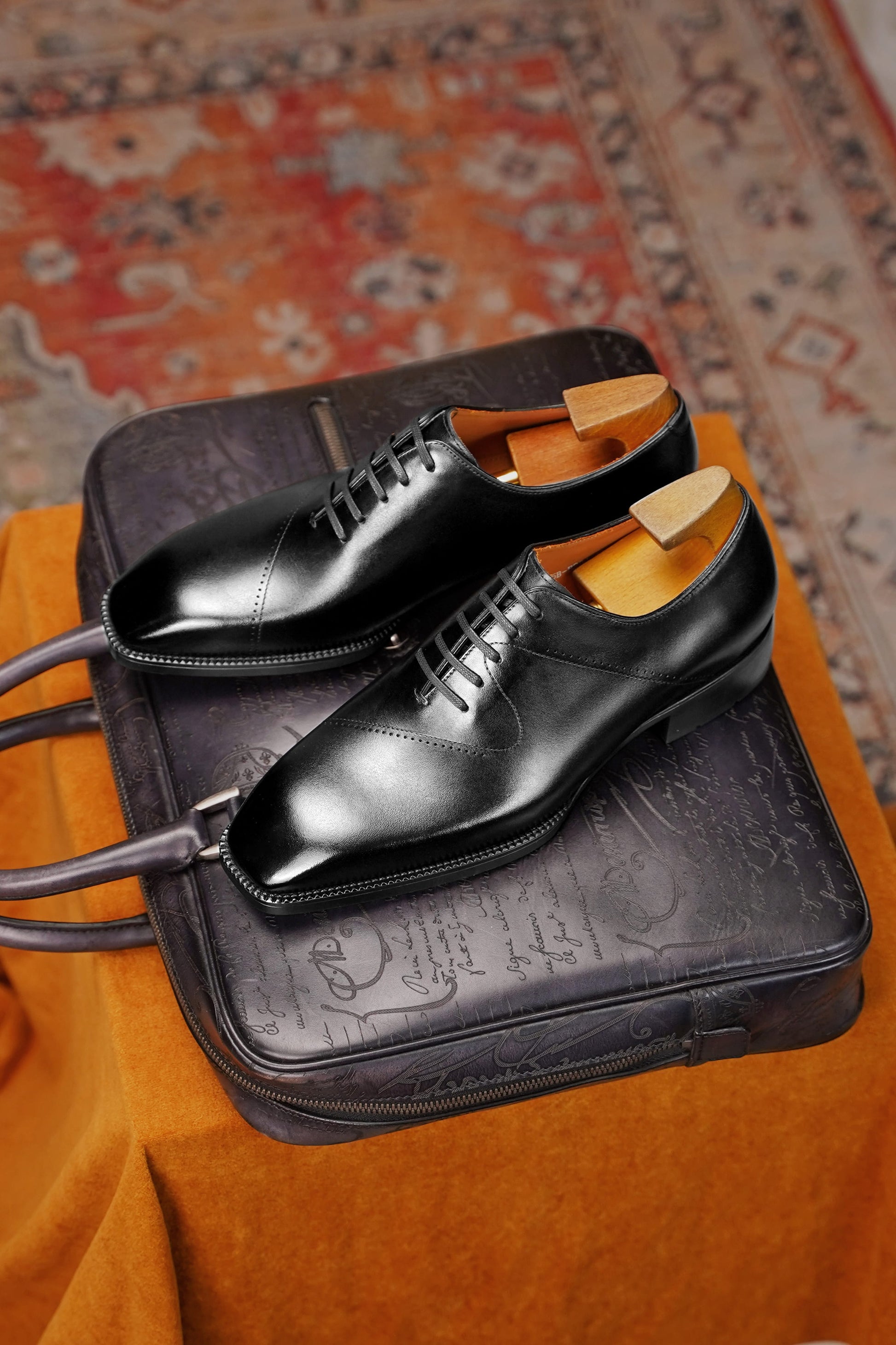A pair of black leather dress shoes with wooden shoe trees, displayed on top of a dark embossed leather case over an orange fabric surface with a patterned rug in the background.
