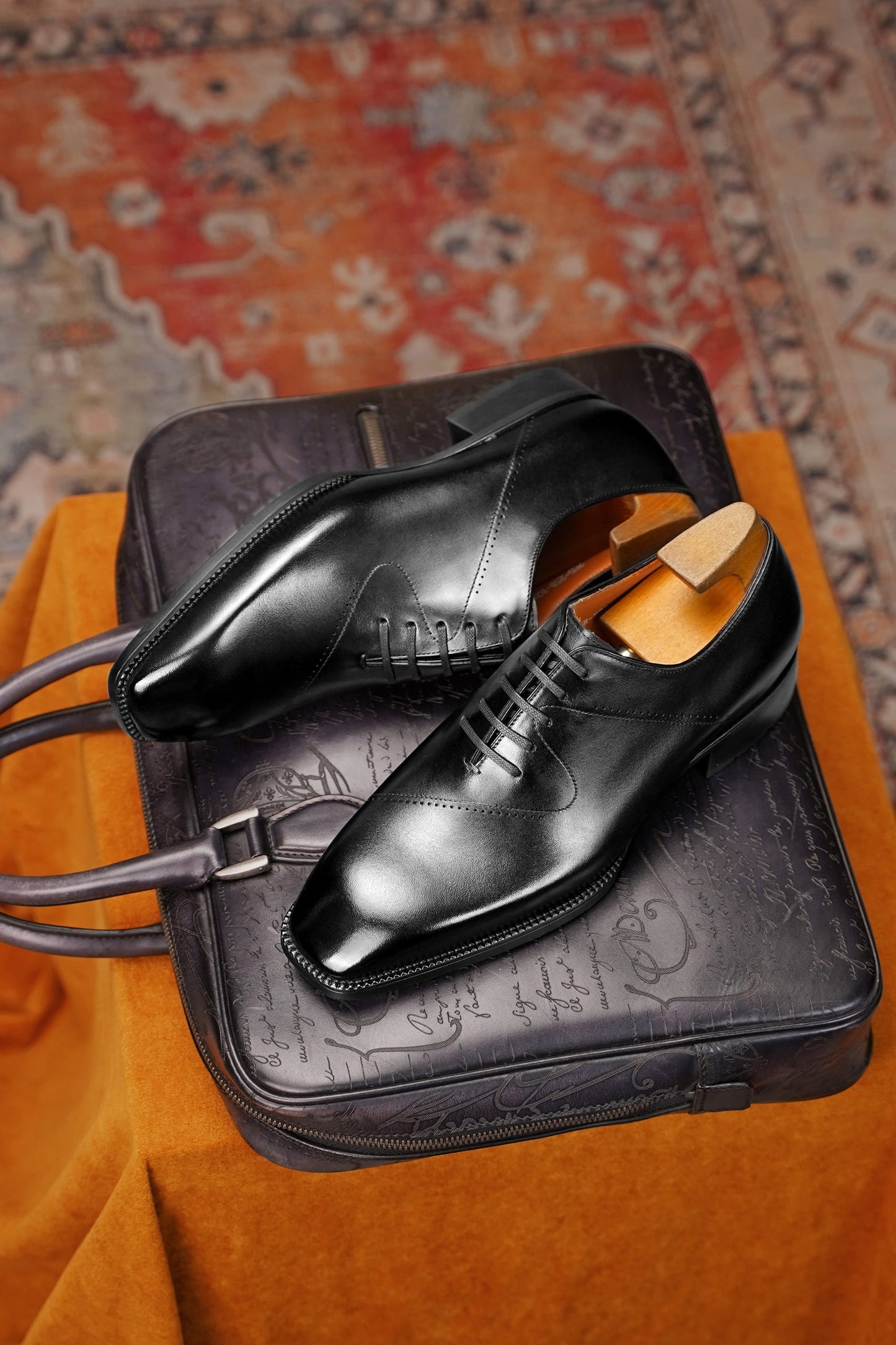 A pair of black leather dress shoes with lace-up fronts and polished finishes, displayed on top of a black embossed leather briefcase. The scene is styled on an orange fabric surface with a patterned rug in the background.