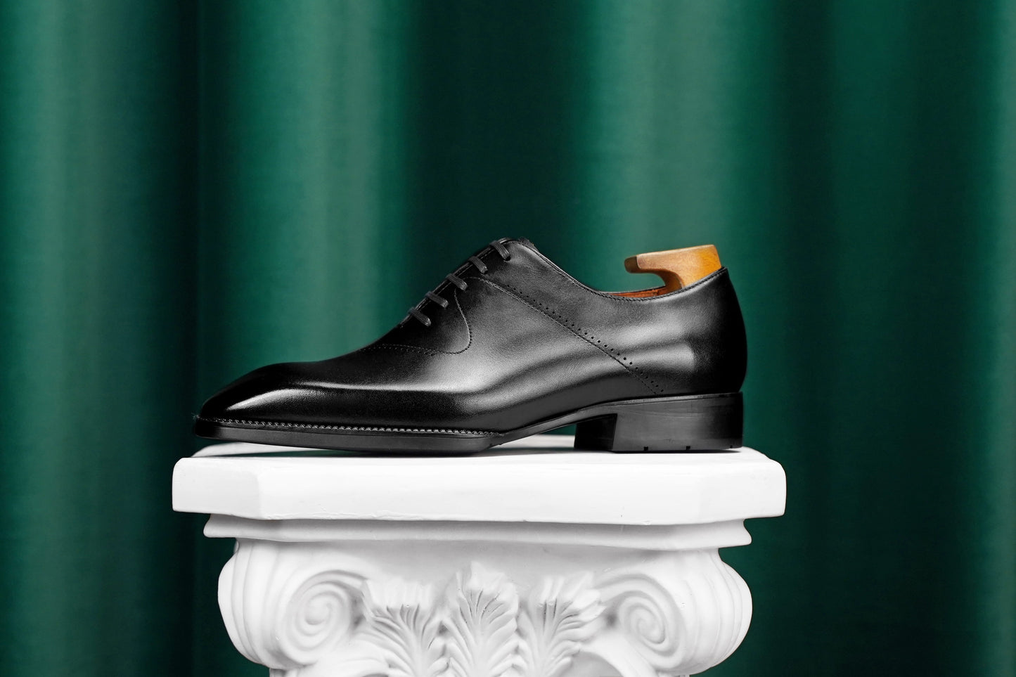 A black leather oxford dress shoe with a low block heel and lace-up front, displayed on a white decorative pedestal against a dark green curtain background.