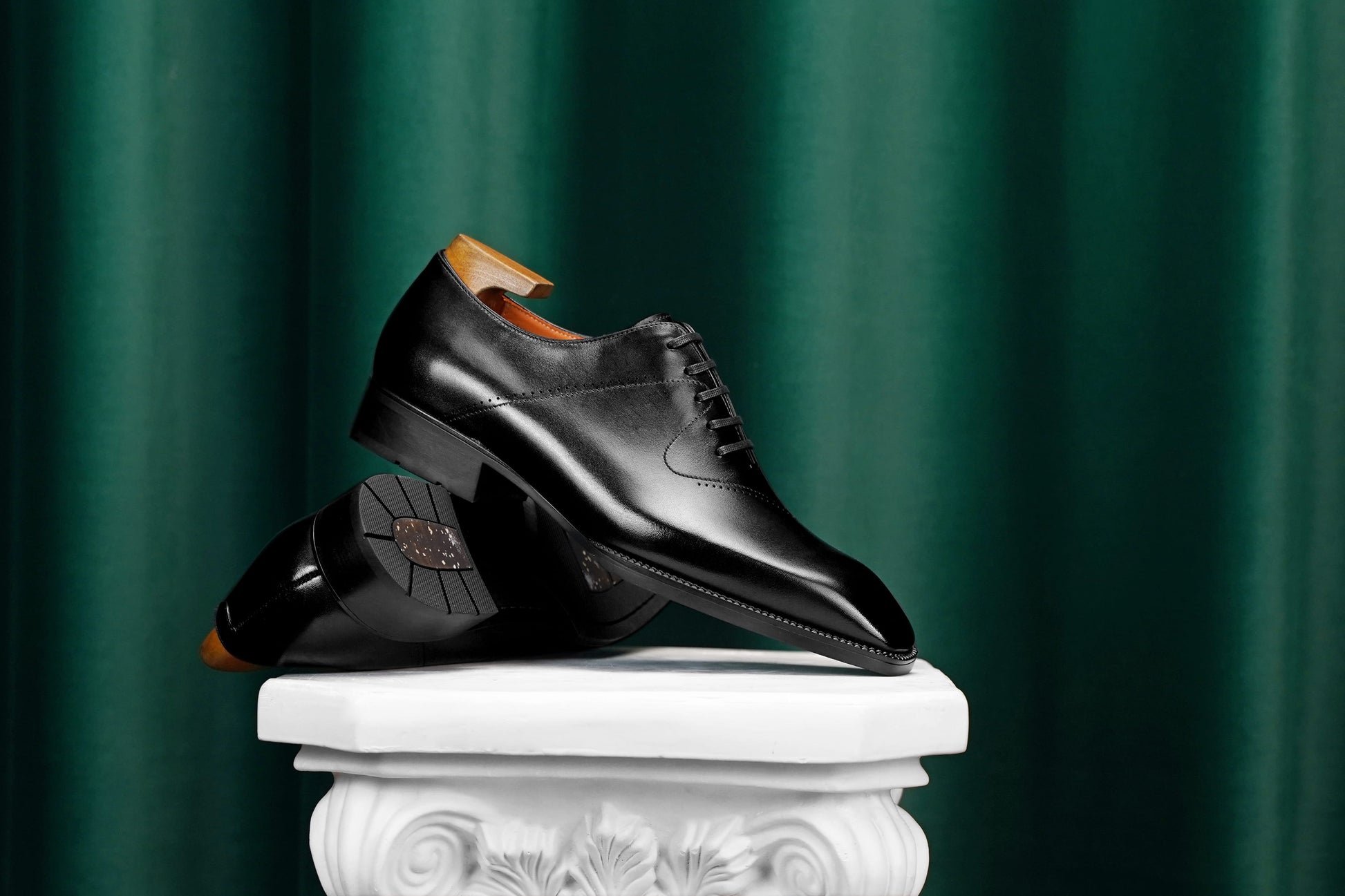A pair of black leather dress shoes displayed on a white decorative pedestal against a dark green curtain background.