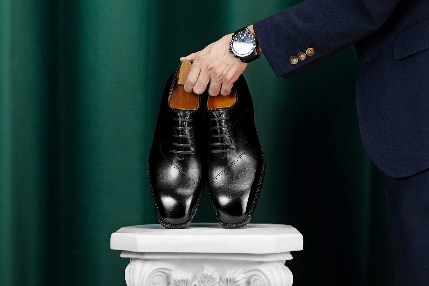 A pair of black leather oxford dress shoes displayed on a white pedestal, with a person's hand holding the shoe trees inside. The person is wearing a dark navy suit and a wristwatch.