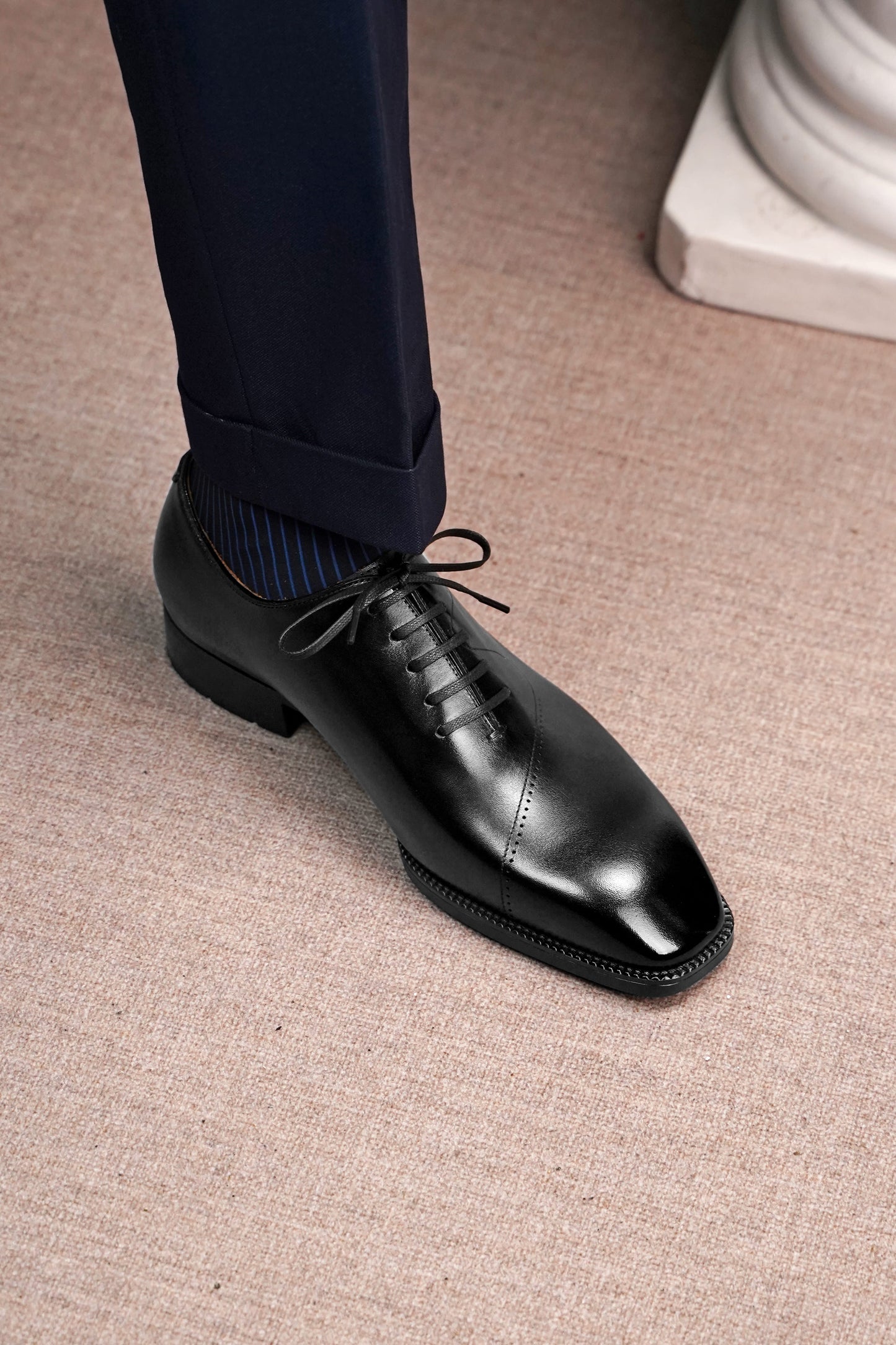A close-up of a man's foot wearing a black leather oxford dress shoe with dark navy trousers and blue striped socks, standing on a textured beige carpet.