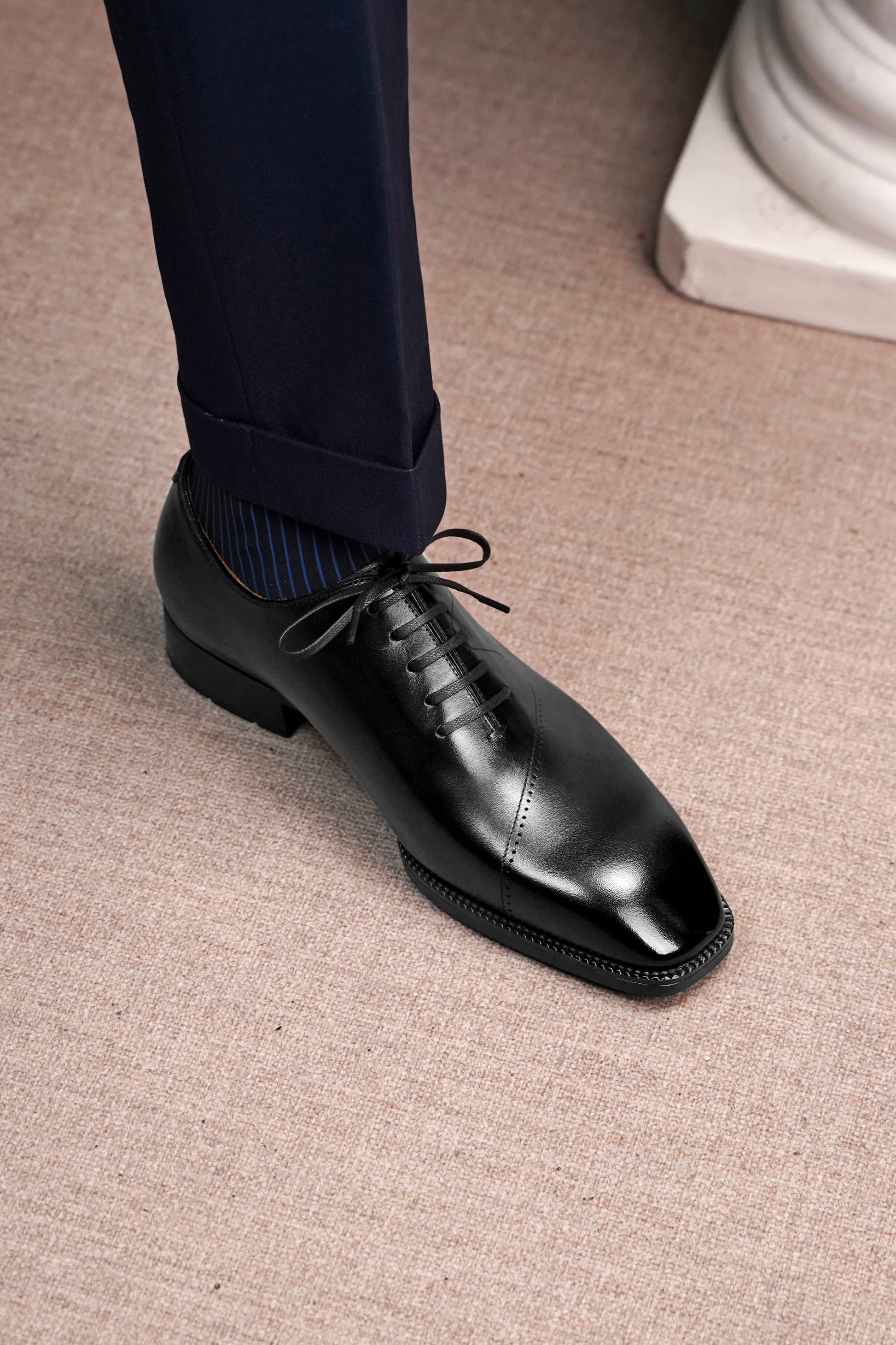 A close-up of a man's foot wearing a black leather oxford dress shoe with dark navy trousers and blue striped socks, standing on a textured beige carpet.
