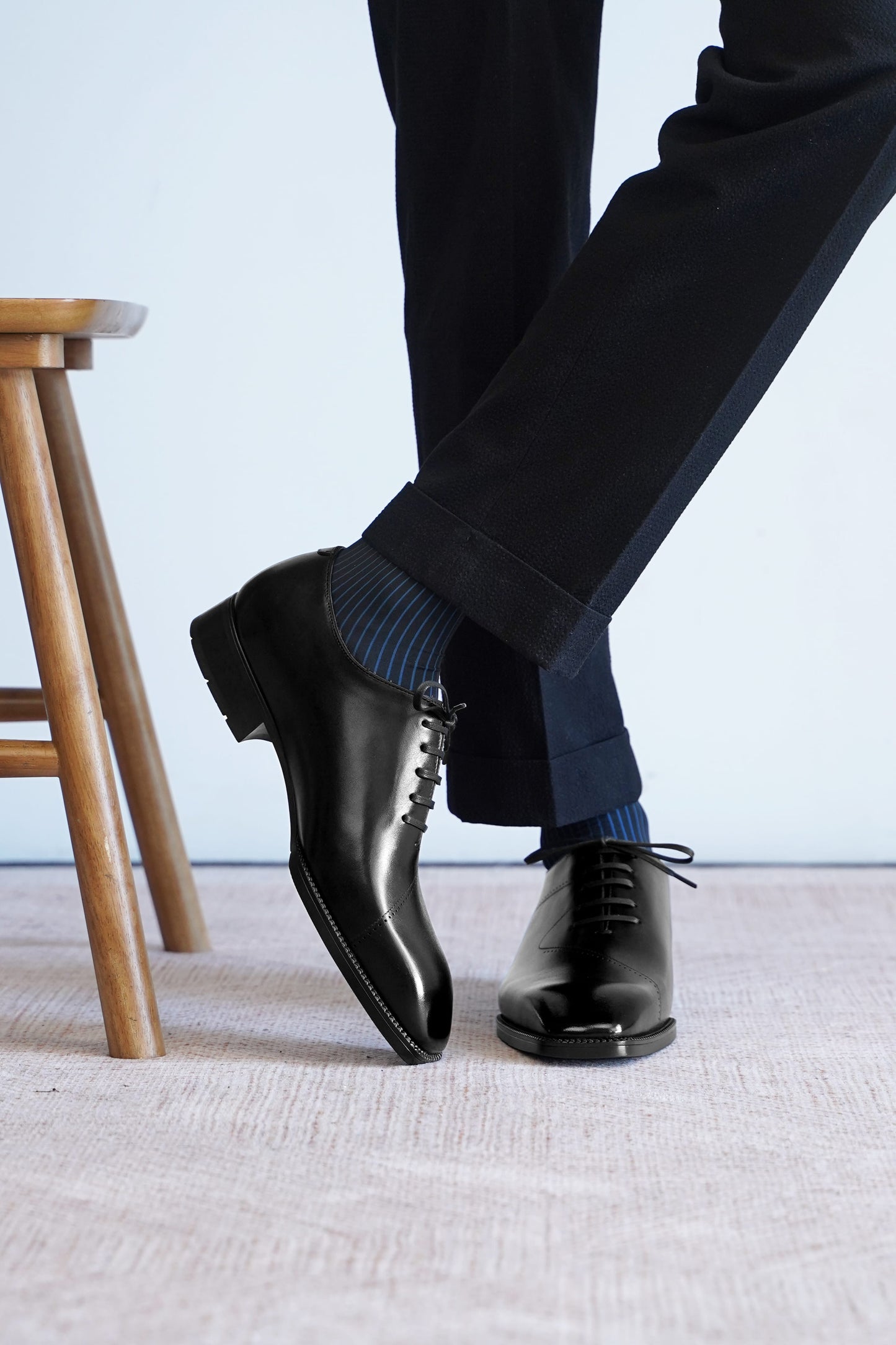 A close-up of a person's legs wearing polished black oxford dress shoes, dark trousers, and blue striped socks, standing on a light textured floor next to a wooden stool.