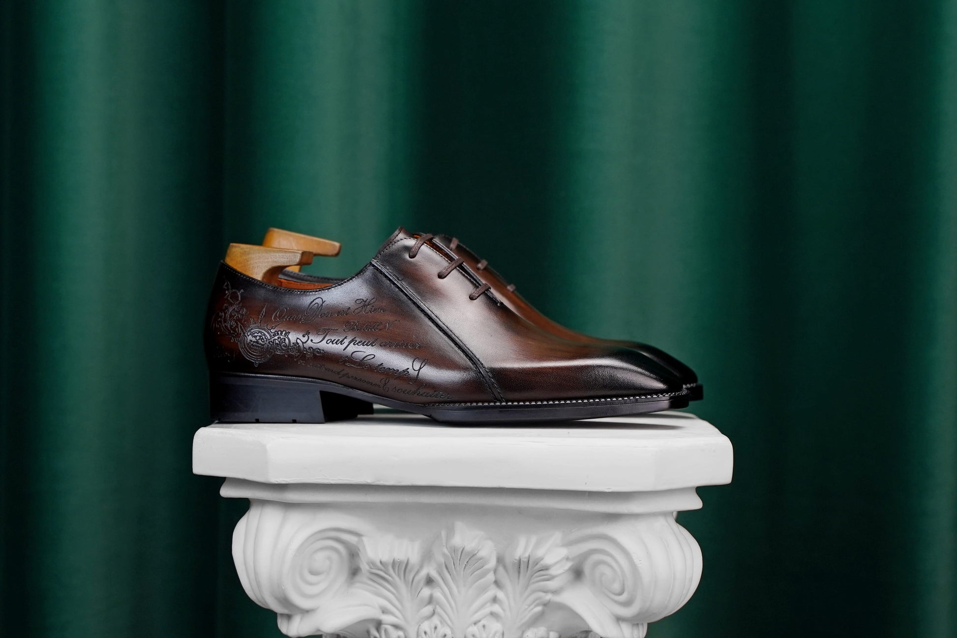 A dark brown leather dress shoe with embossed floral detailing, displayed on a white decorative pedestal against a dark green curtain background.