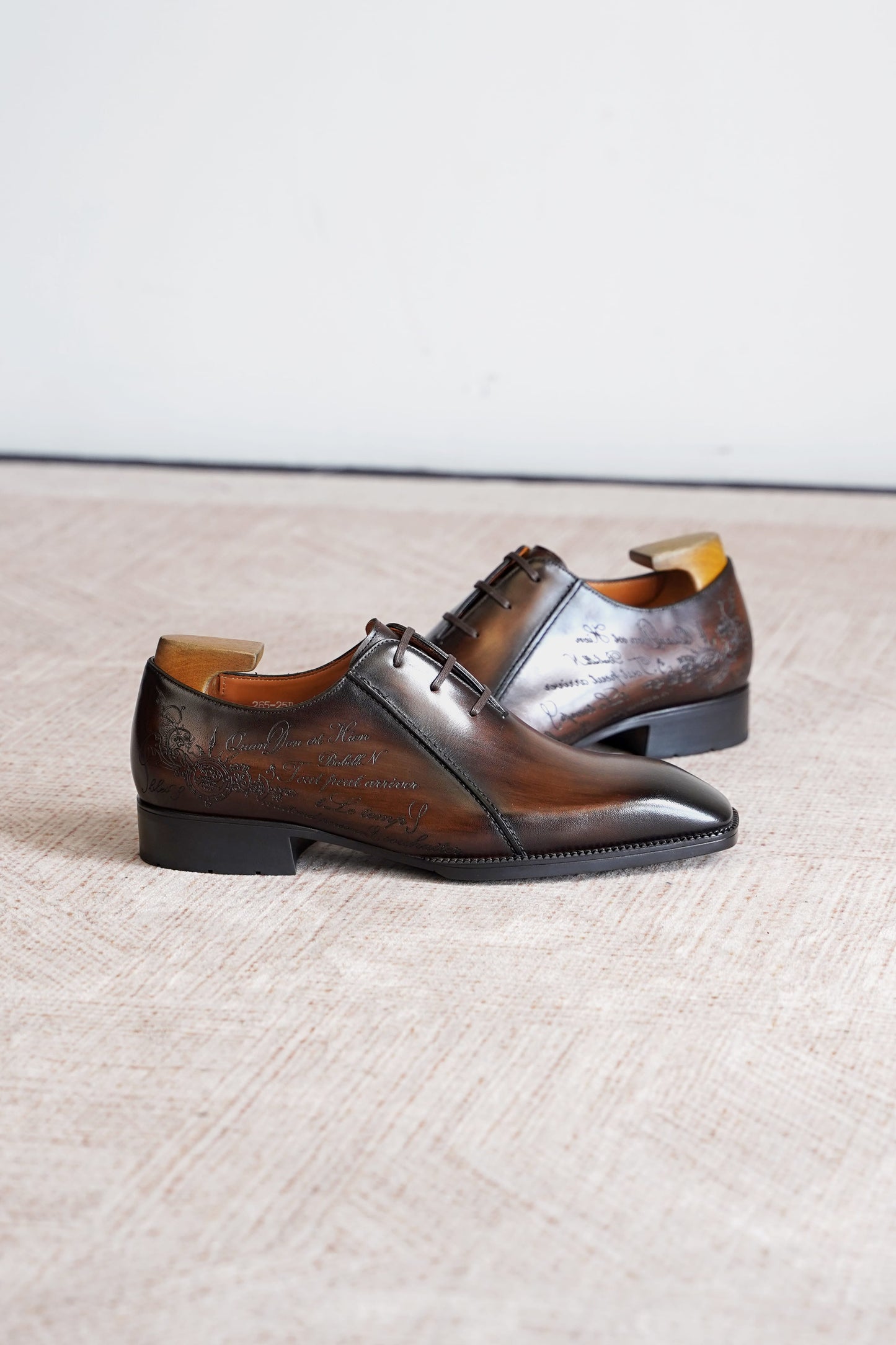 A pair of dark brown leather oxford dress shoes with a glossy finish and subtle embossed detailing, displayed on a textured beige floor against a light gray wall.