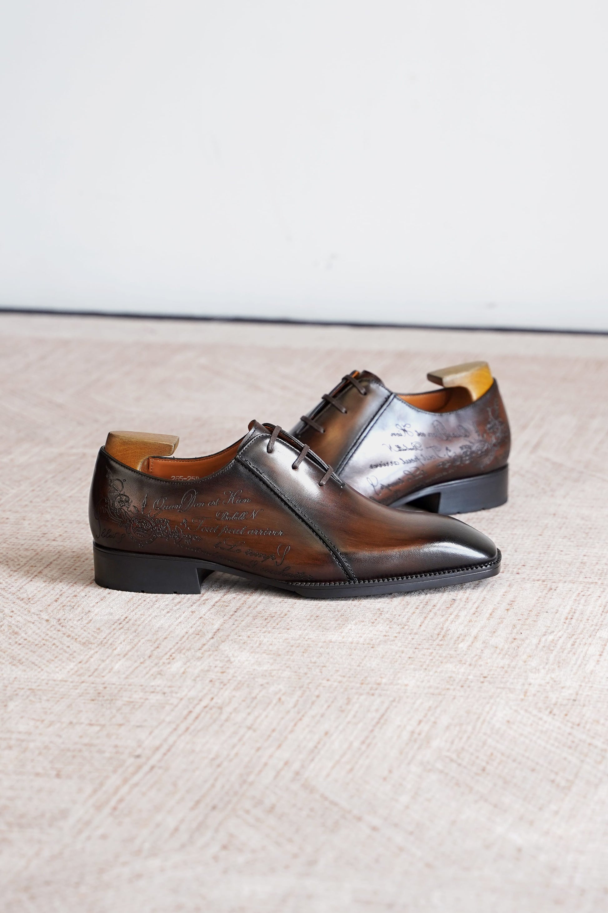 A pair of dark brown leather oxford dress shoes with a glossy finish and subtle embossed detailing, displayed on a textured beige floor against a light gray wall.