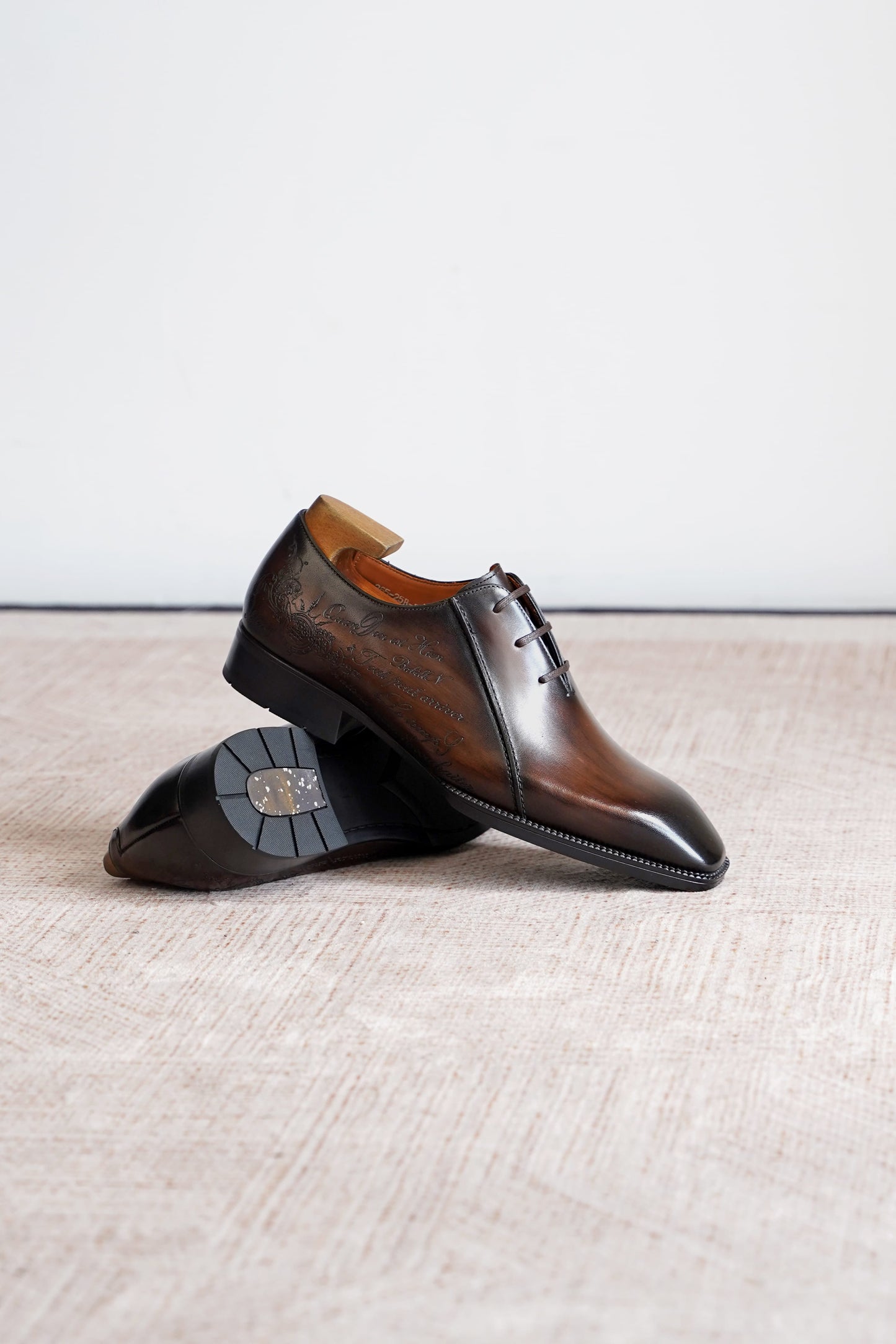 A pair of men's brown leather oxford shoes with a glossy finish and decorative stitching, displayed on a textured beige surface against a plain white background.