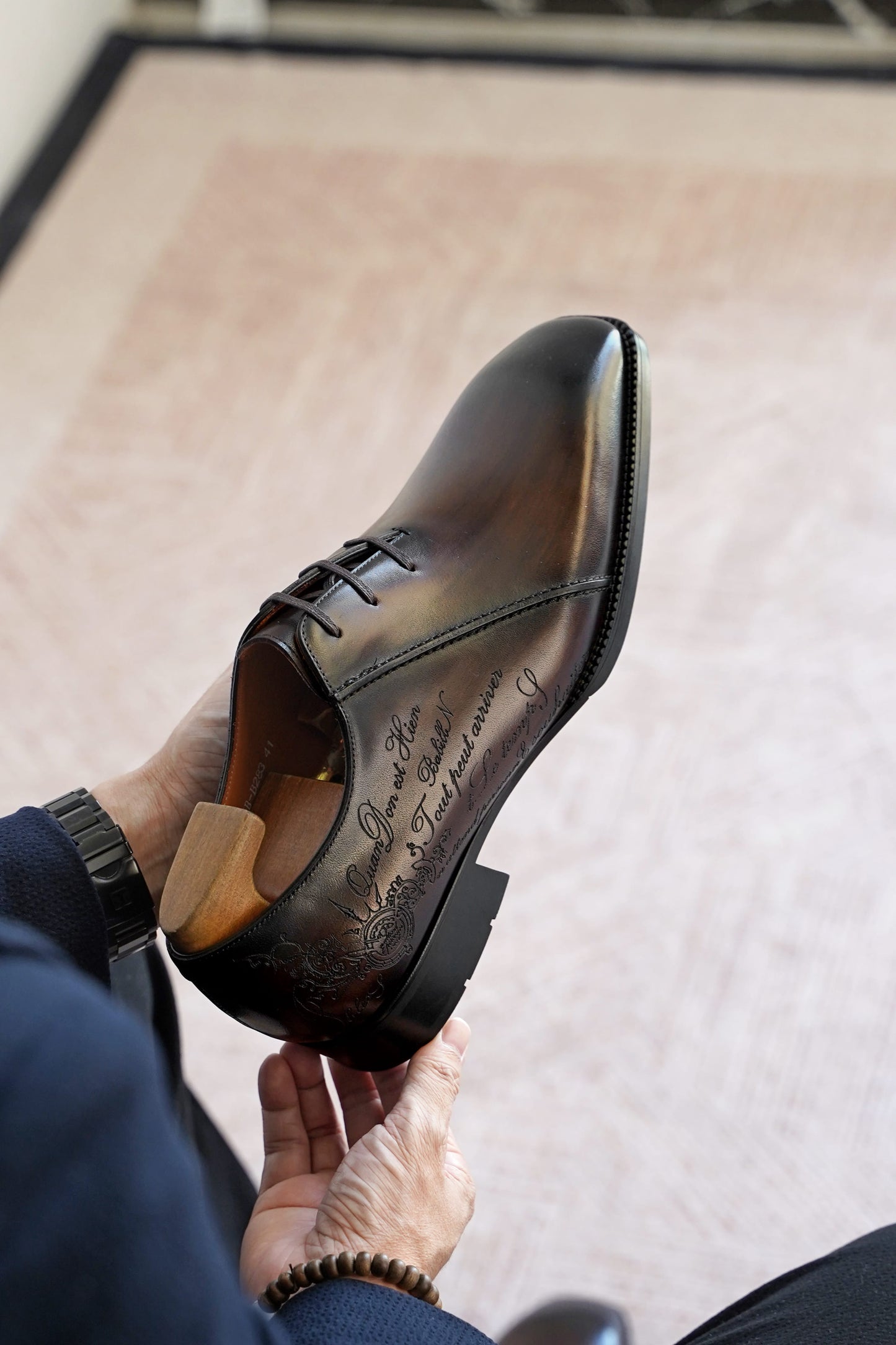 A person holds a polished dark brown leather dress shoe with hand-lettered script on the side, shown from a high angle against a light beige rug.