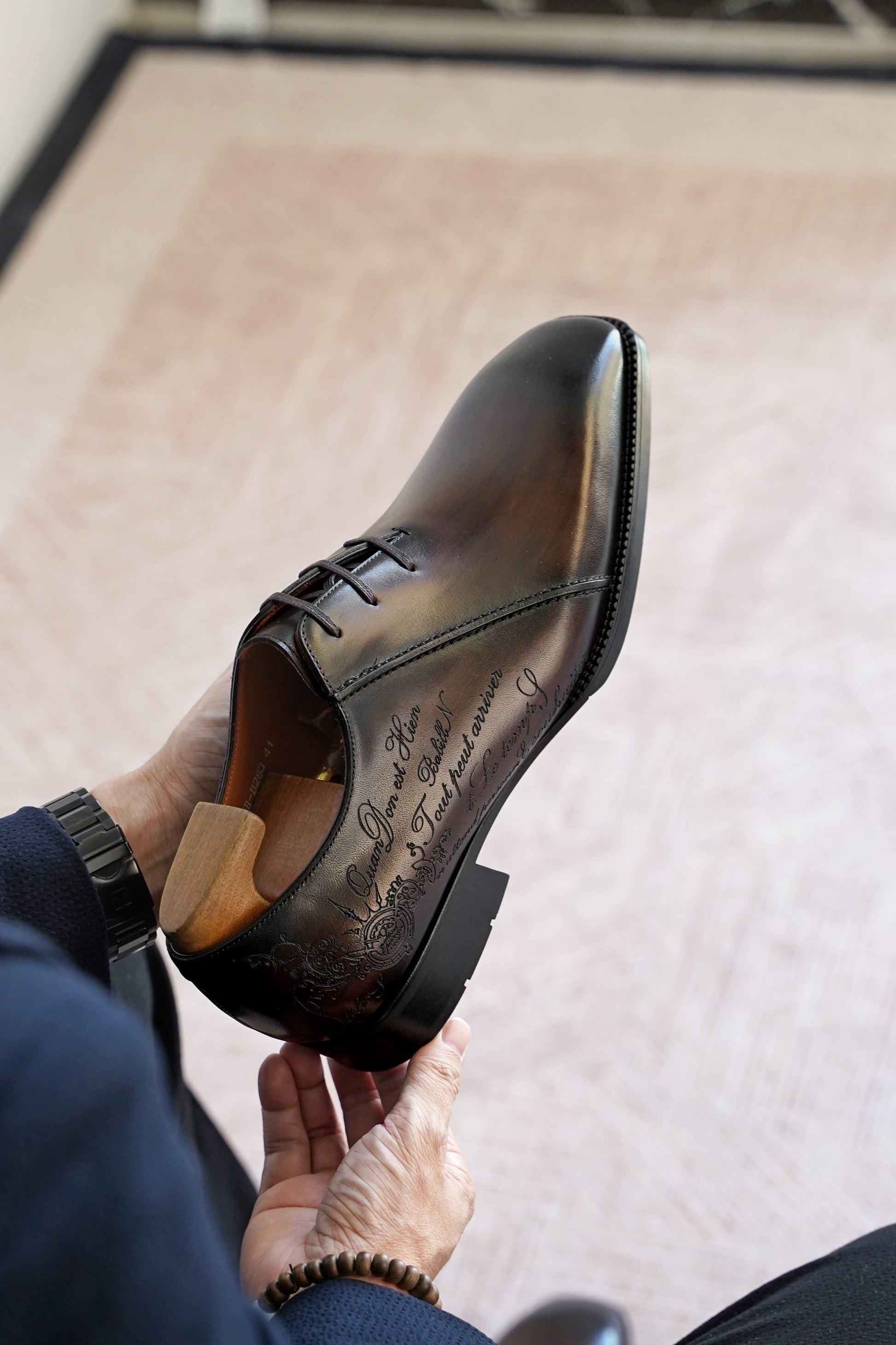 A person holds a polished dark brown leather dress shoe with hand-lettered script on the side, shown from a high angle against a light beige rug.