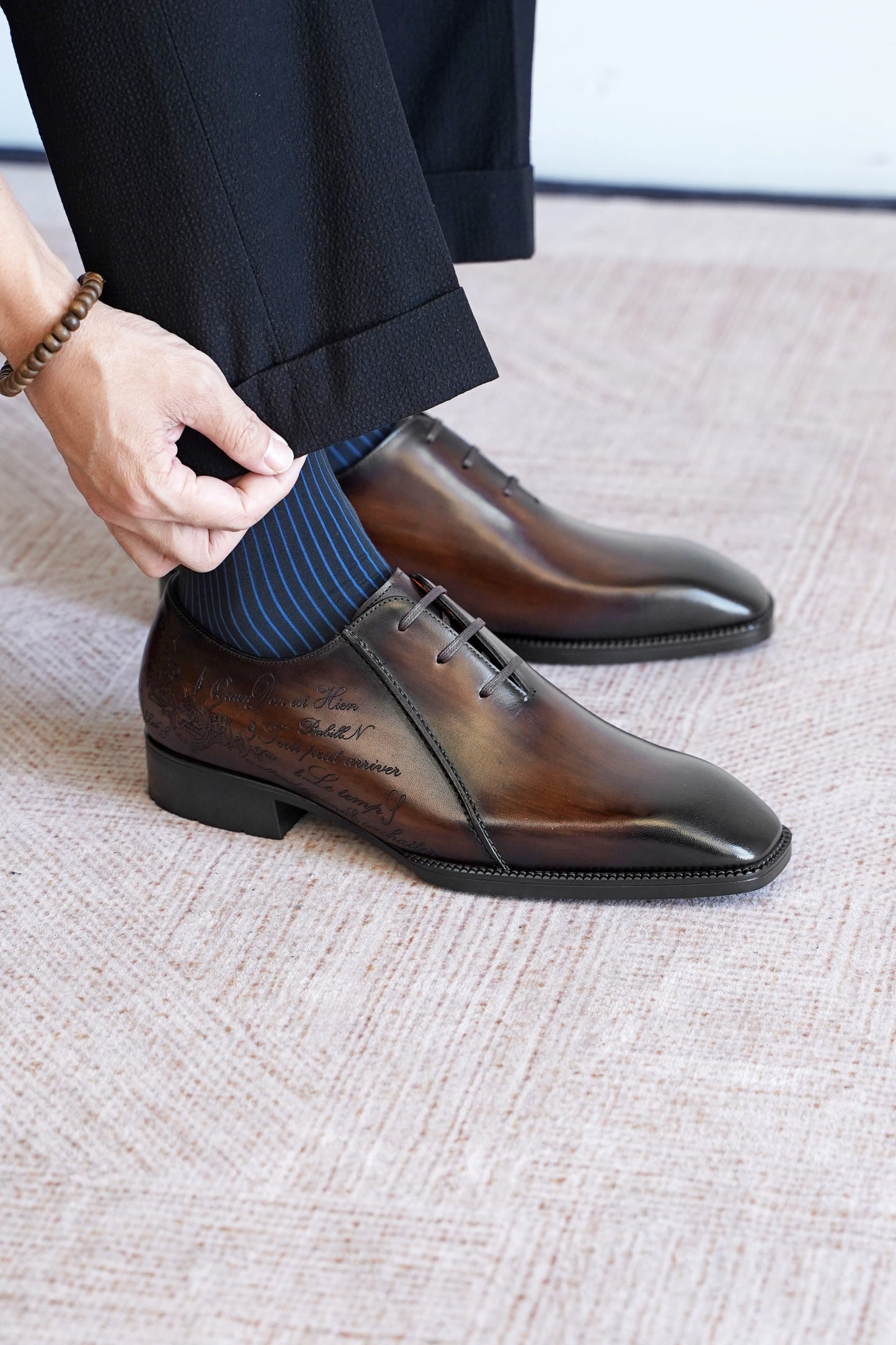 A close-up of a person wearing polished dark brown oxford dress shoes with a striped blue sock, paired with black trousers, standing on a textured beige carpet.