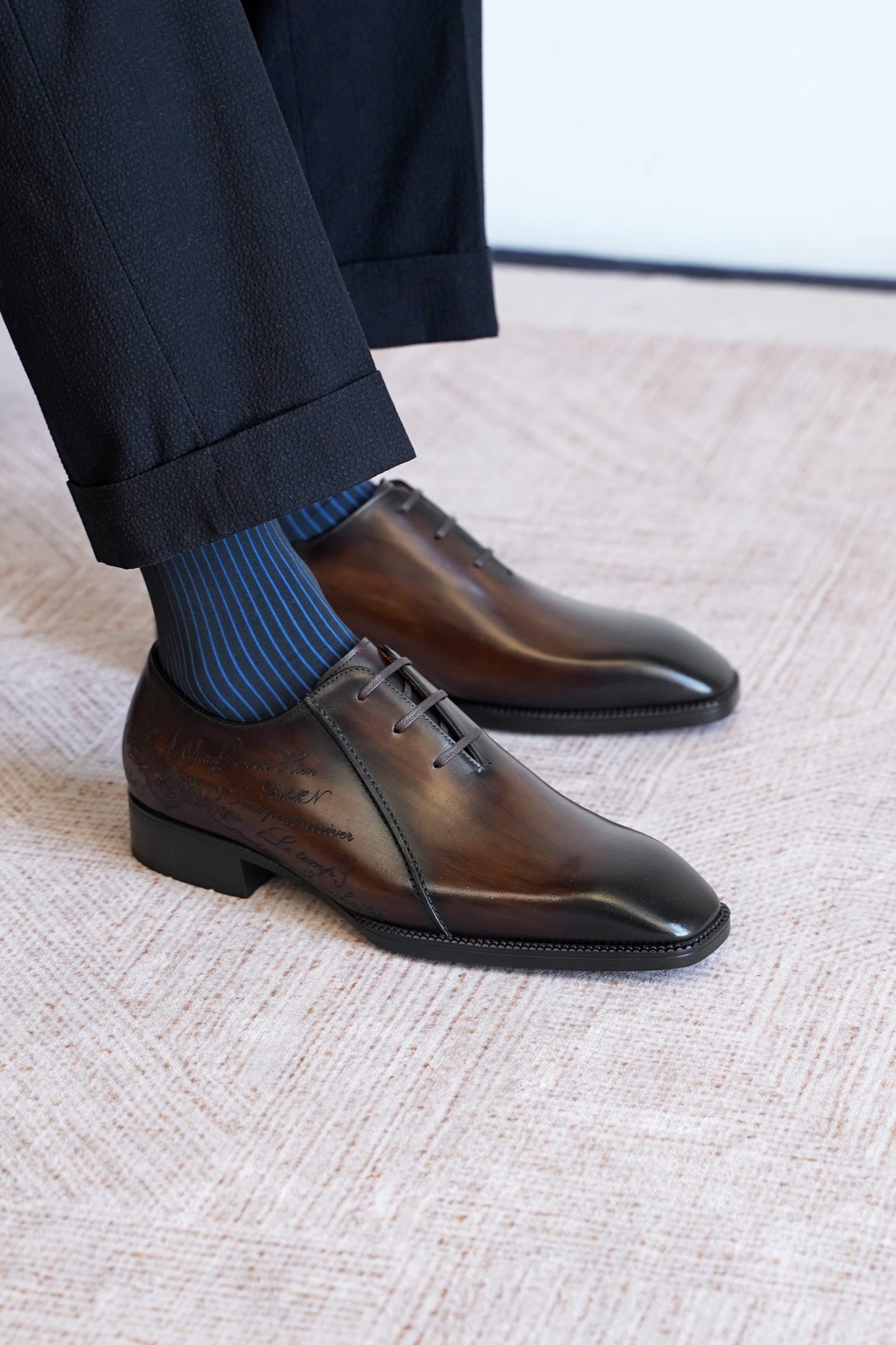 A close-up of a person's feet wearing dark brown oxford dress shoes with a glossy finish, paired with blue striped socks and dark trousers, standing on a textured beige carpet.