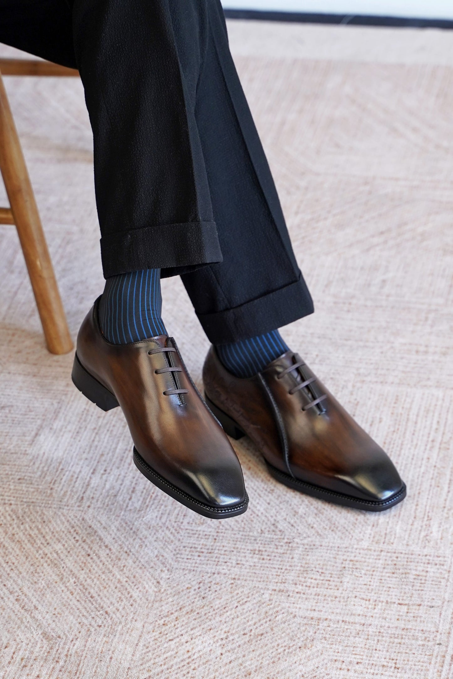 A close-up of a person's feet wearing dark brown oxford dress shoes with a glossy finish, paired with dark blue socks and black trousers, sitting on a textured beige carpet.