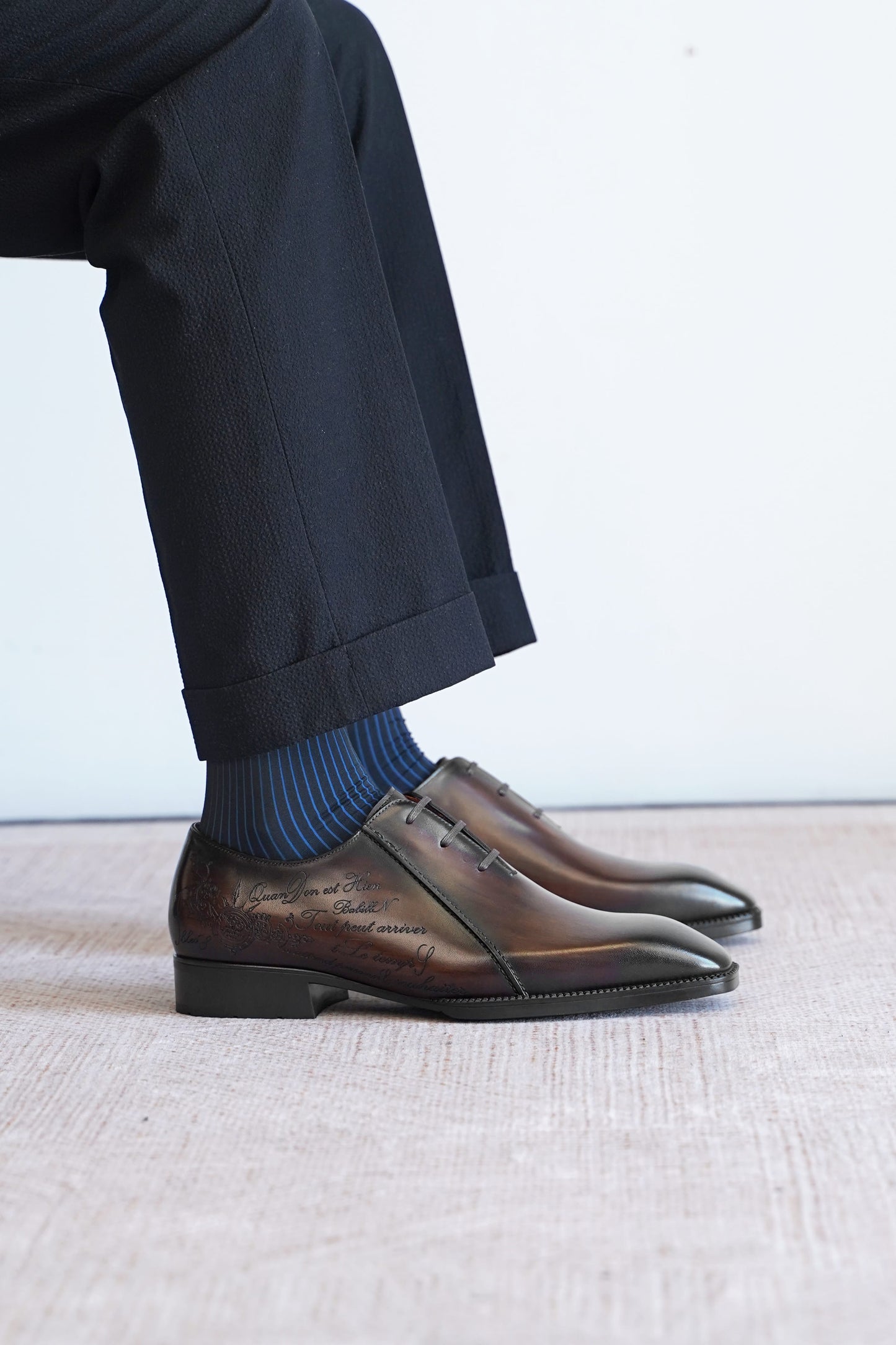 A close-up of a person's legs wearing dark brown embossed leather oxford shoes, blue striped socks, and dark trousers, sitting on a light textured floor against a white wall.