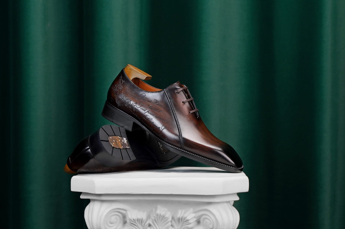 A pair of dark brown leather dress shoes with a glossy finish and subtle embossed detailing, displayed on a white classical-style pedestal against a dark green draped background.