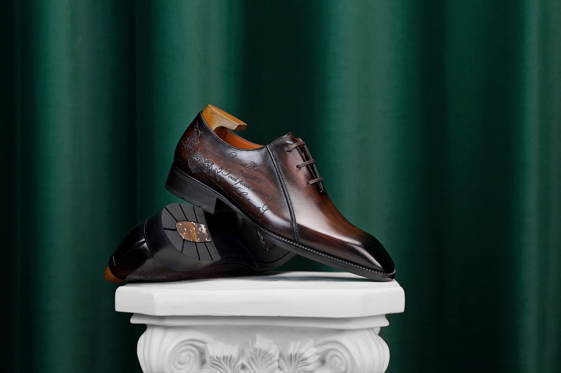 A pair of dark brown leather dress shoes with a glossy finish and subtle embossed detailing, displayed on a white classical-style pedestal against a dark green draped background.