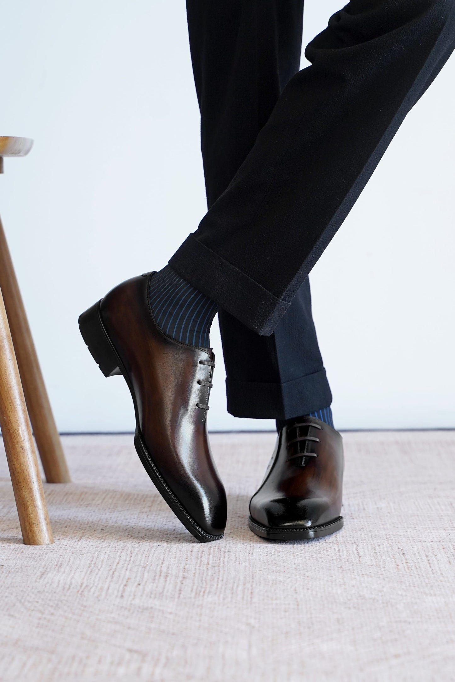 A person wearing dark brown oxford dress shoes with dark blue striped socks and black trousers, standing on a light beige textured rug.