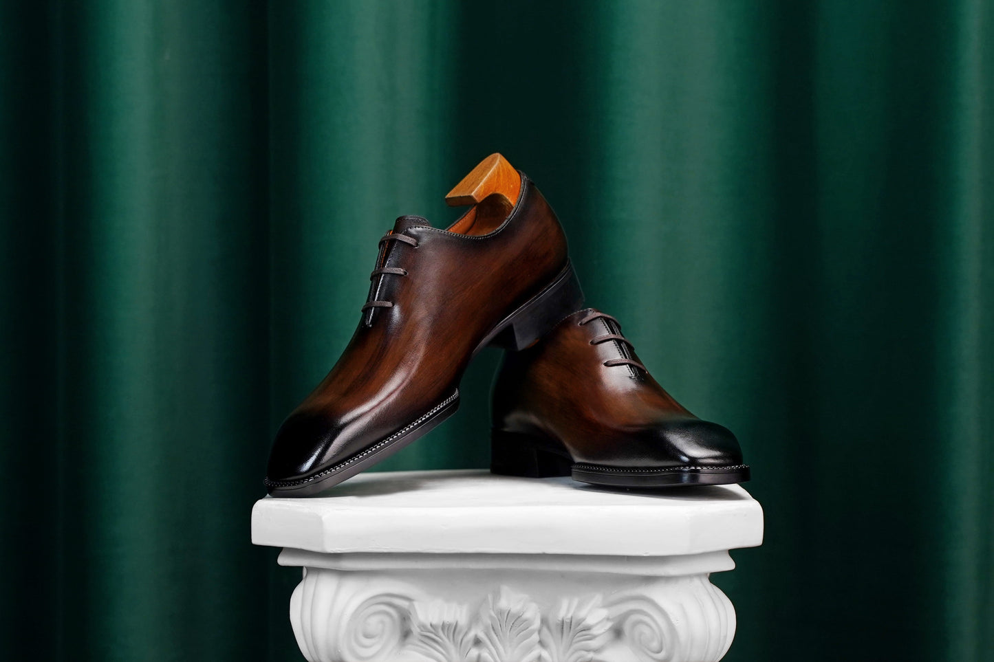 A pair of polished dark brown leather oxford dress shoes with black toe caps, displayed on a white decorative pedestal against a dark green curtain backdrop.