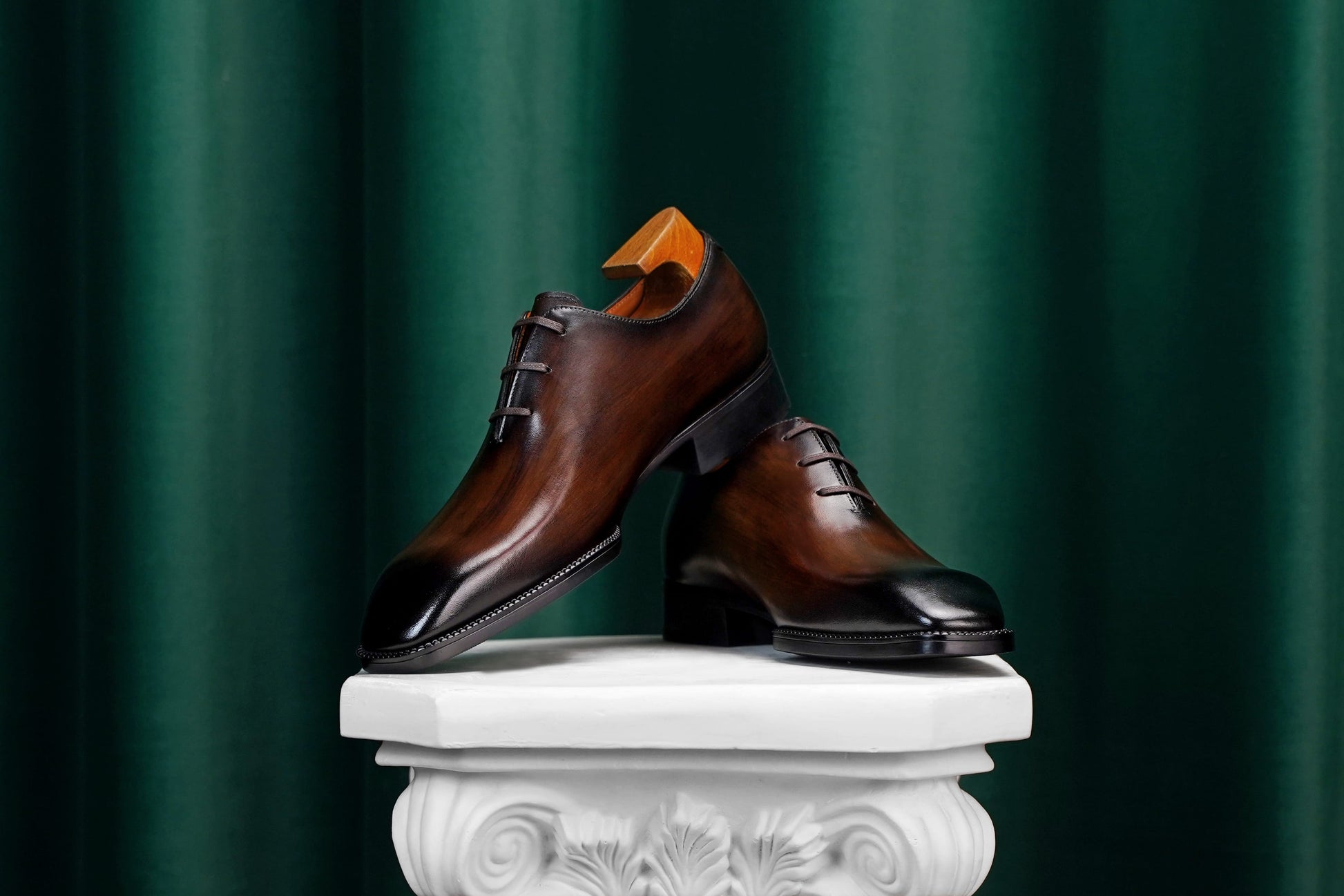 A pair of polished dark brown leather oxford dress shoes with black toe caps, displayed on a white decorative pedestal against a dark green curtain backdrop.