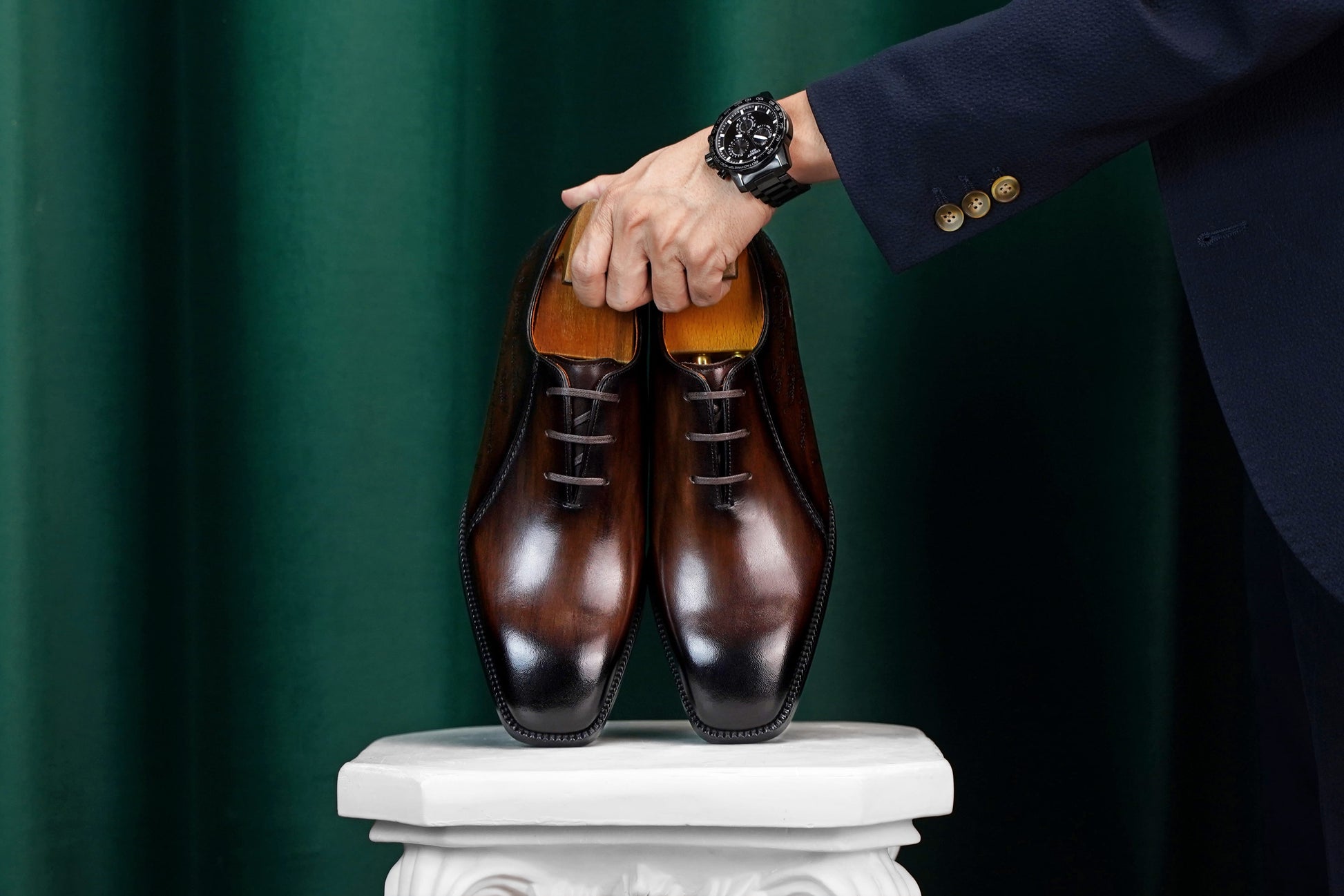 A pair of polished dark brown oxford dress shoes with a glossy finish, held by a person wearing a navy blazer and a black watch, set against a dark green curtain backdrop.