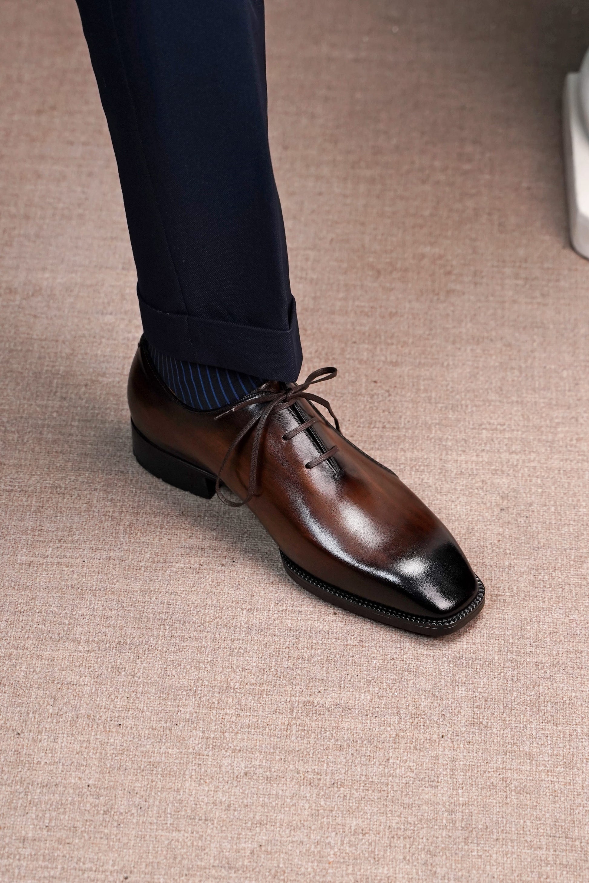 A close-up of a man's foot wearing a polished dark brown oxford dress shoe with a black pointed toe, paired with navy trousers and blue striped socks, standing on a textured beige carpet.