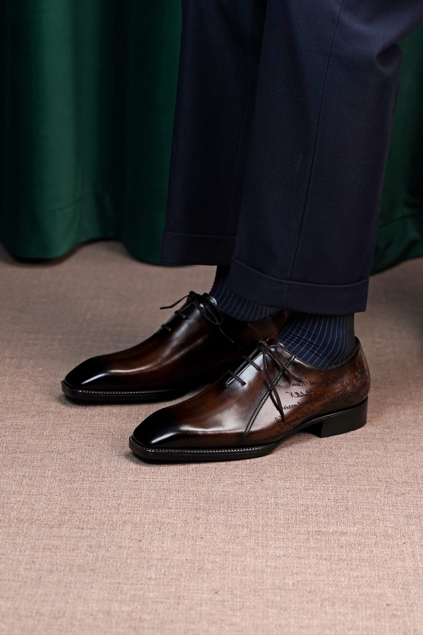 A close-up of a person wearing dark brown polished oxford dress shoes with dark blue socks and navy trousers, standing on a beige textured floor in front of a dark green curtain.