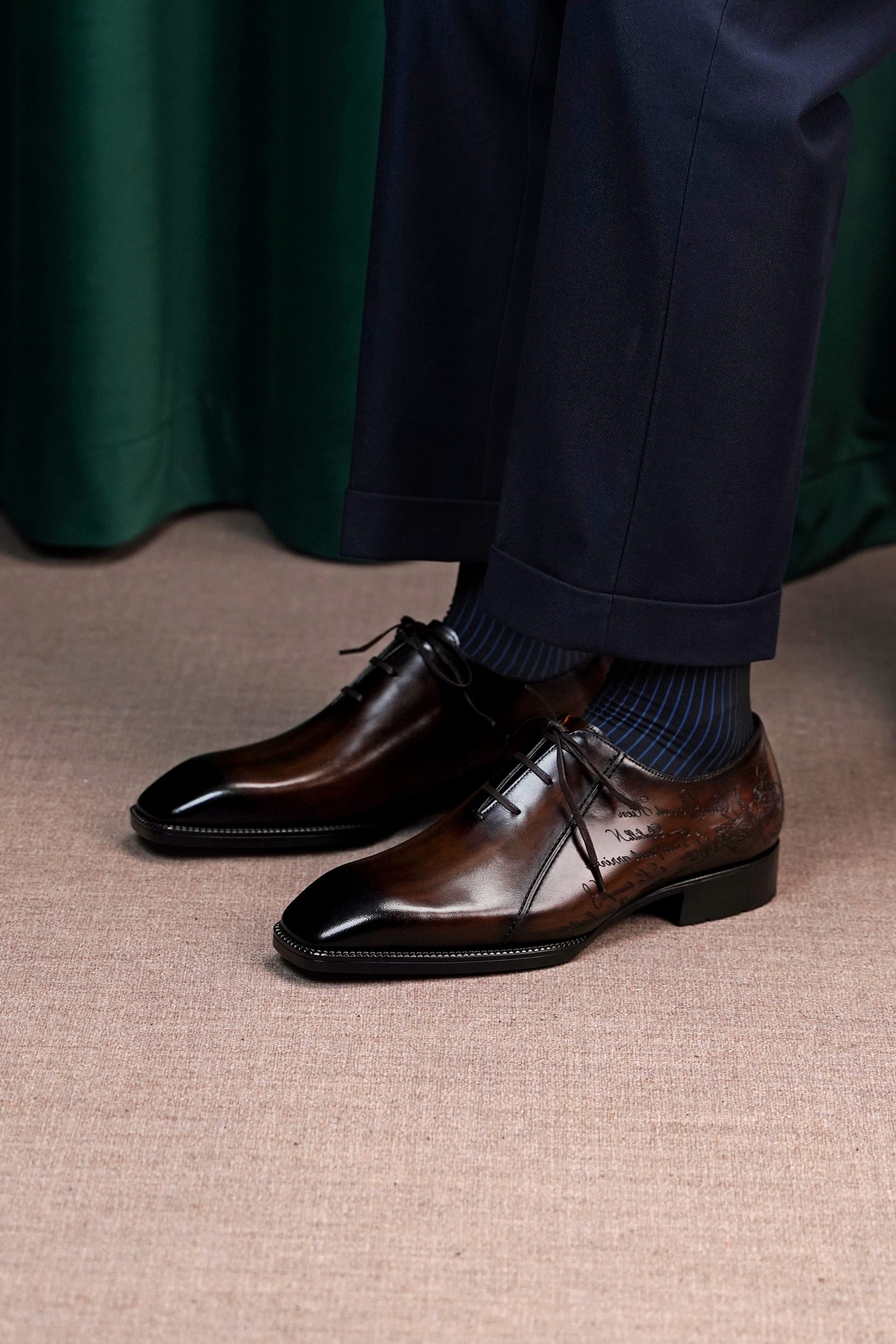 A close-up of a person wearing dark brown polished oxford dress shoes with dark blue socks and navy trousers, standing on a beige textured floor in front of a dark green curtain.