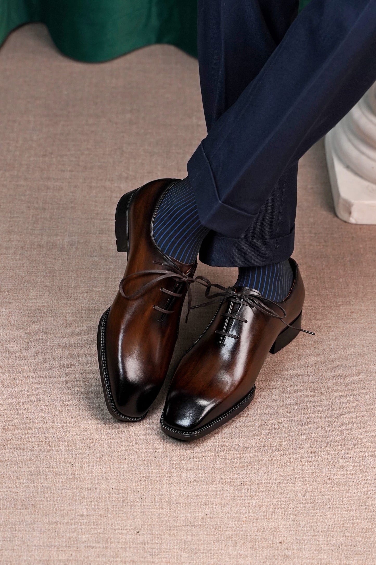 A pair of polished dark brown oxford dress shoes with a glossy finish, worn with navy blue dress pants and blue striped socks on a textured beige carpet.