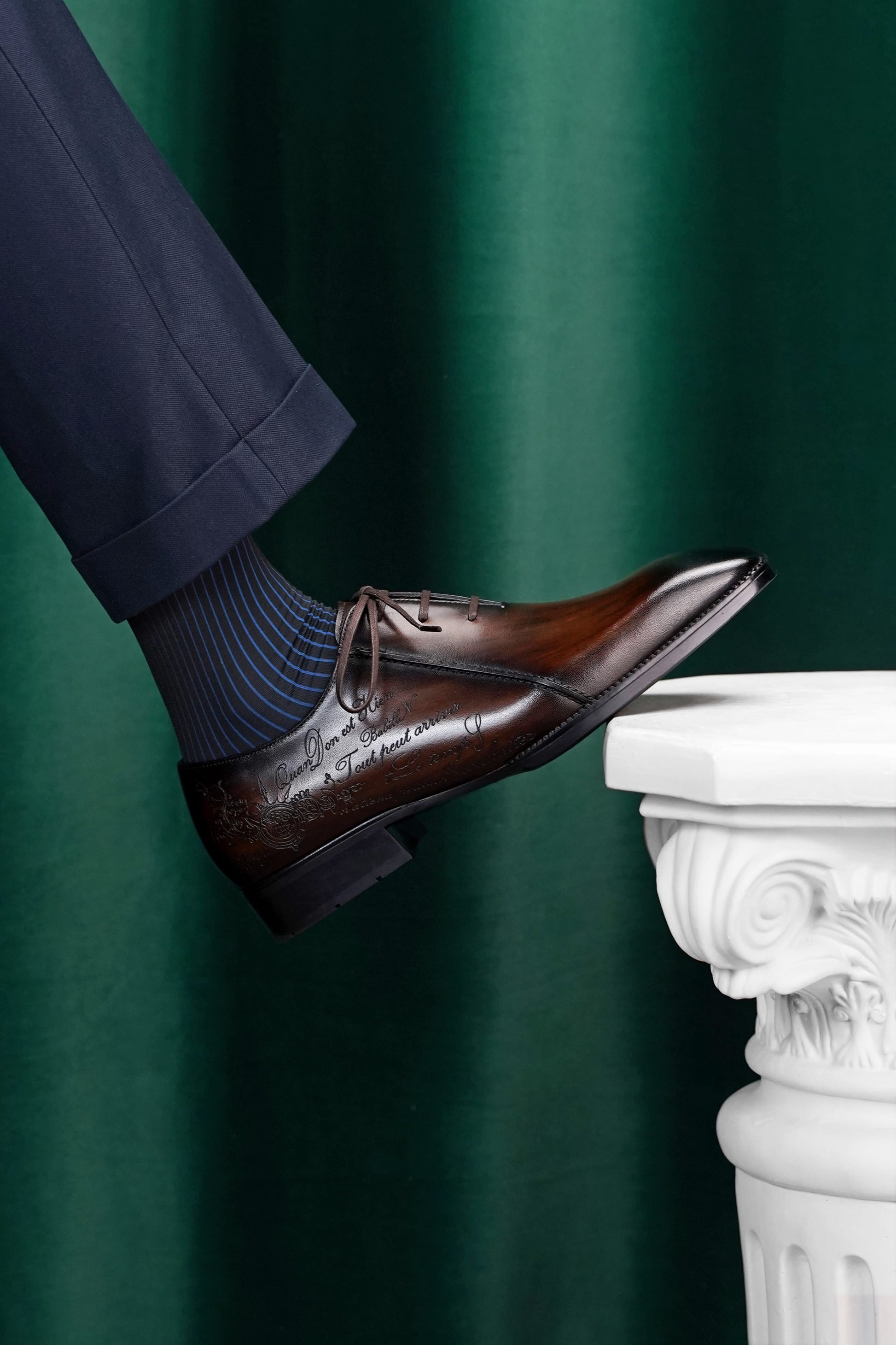 A close-up of a person's foot wearing a dark brown oxford dress shoe with dark blue striped socks, resting on the edge of a white classical-style pedestal against a dark green curtain background.