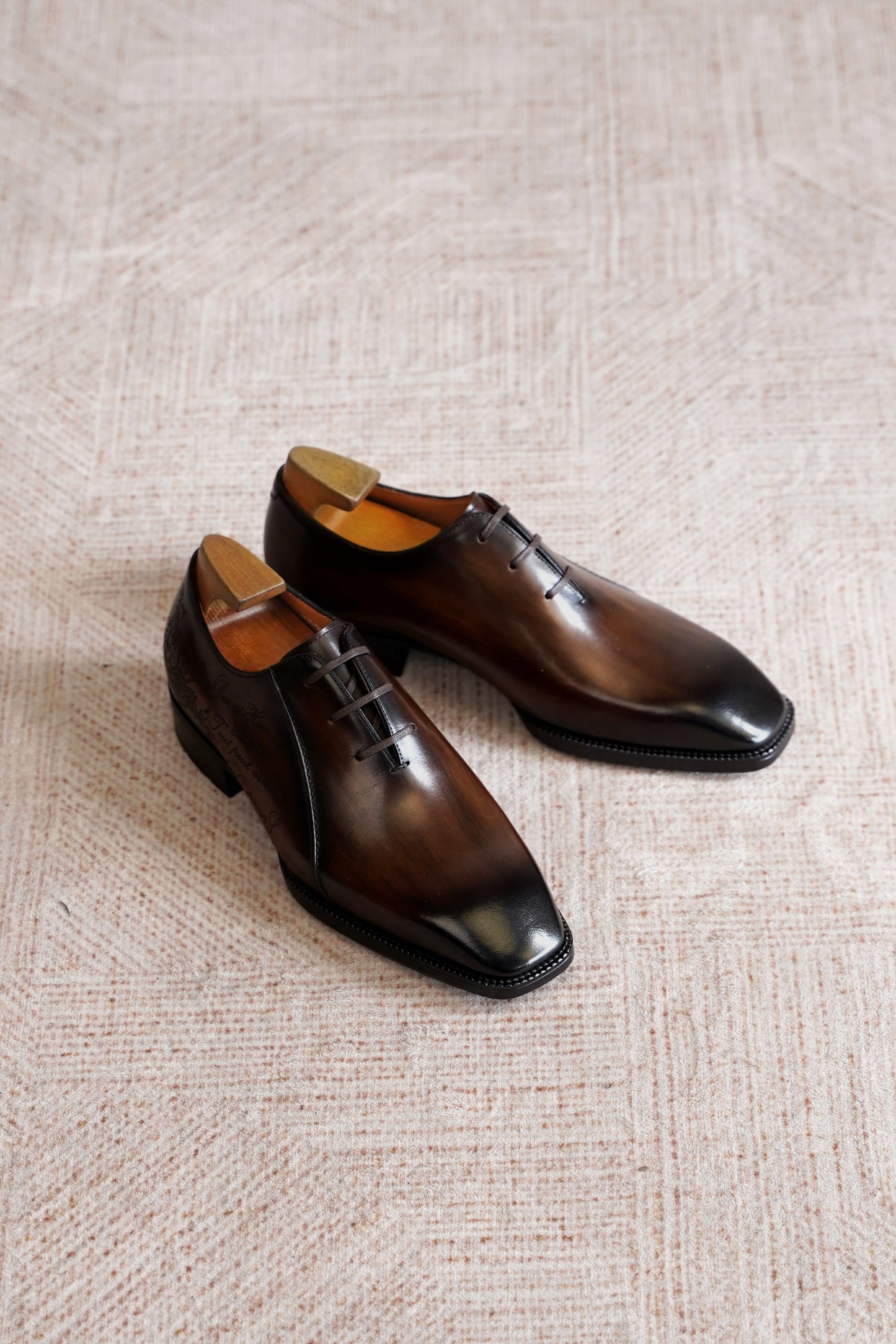 A pair of dark brown leather oxford dress shoes with a glossy finish, displayed on a textured beige woven rug.