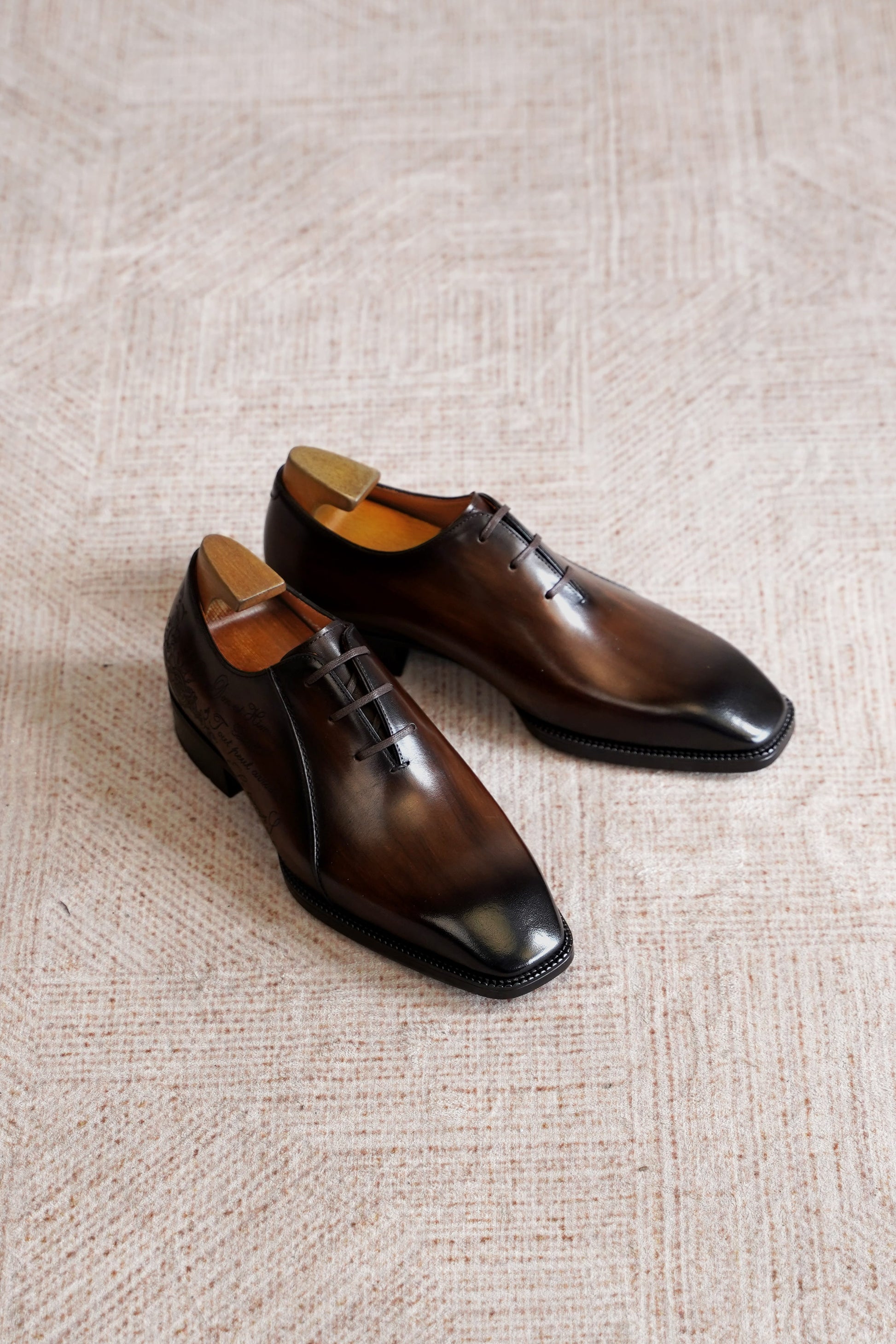 A pair of dark brown leather oxford dress shoes with a glossy finish, displayed on a textured beige woven rug.