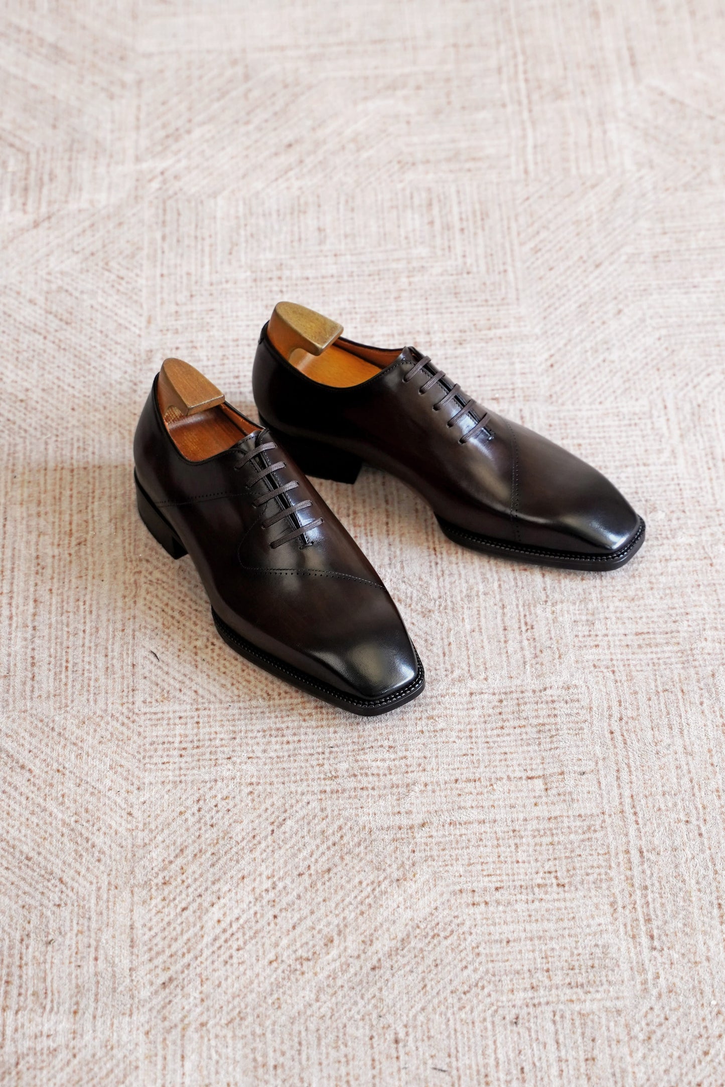 A pair of dark brown leather oxford dress shoes with a square toe, displayed on a textured beige woven rug.