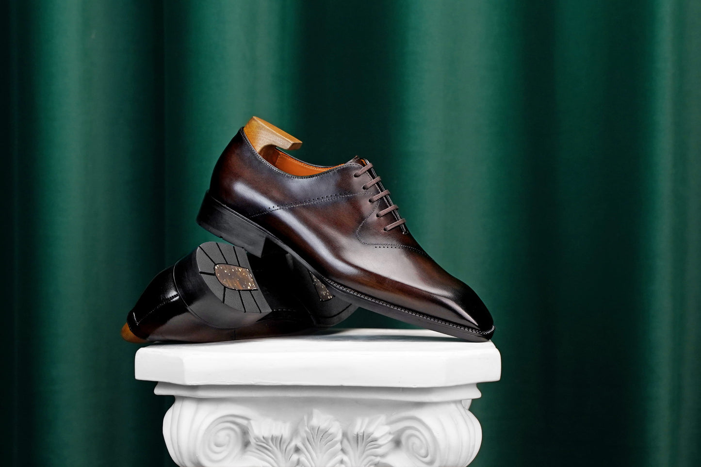 A pair of dark brown leather dress shoes with a polished finish and lace-up closure, displayed on a white decorative pedestal against a dark green curtain background.