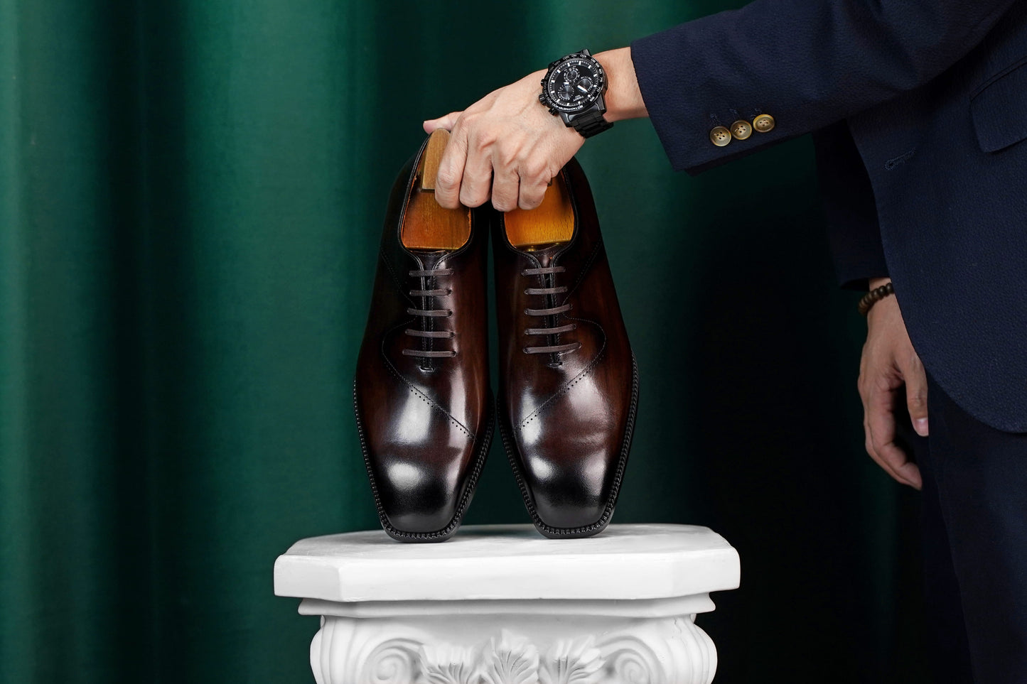 A pair of dark brown oxford dress shoes displayed on a white pedestal, with a person in a navy blazer holding the heels against a dark green curtain background.