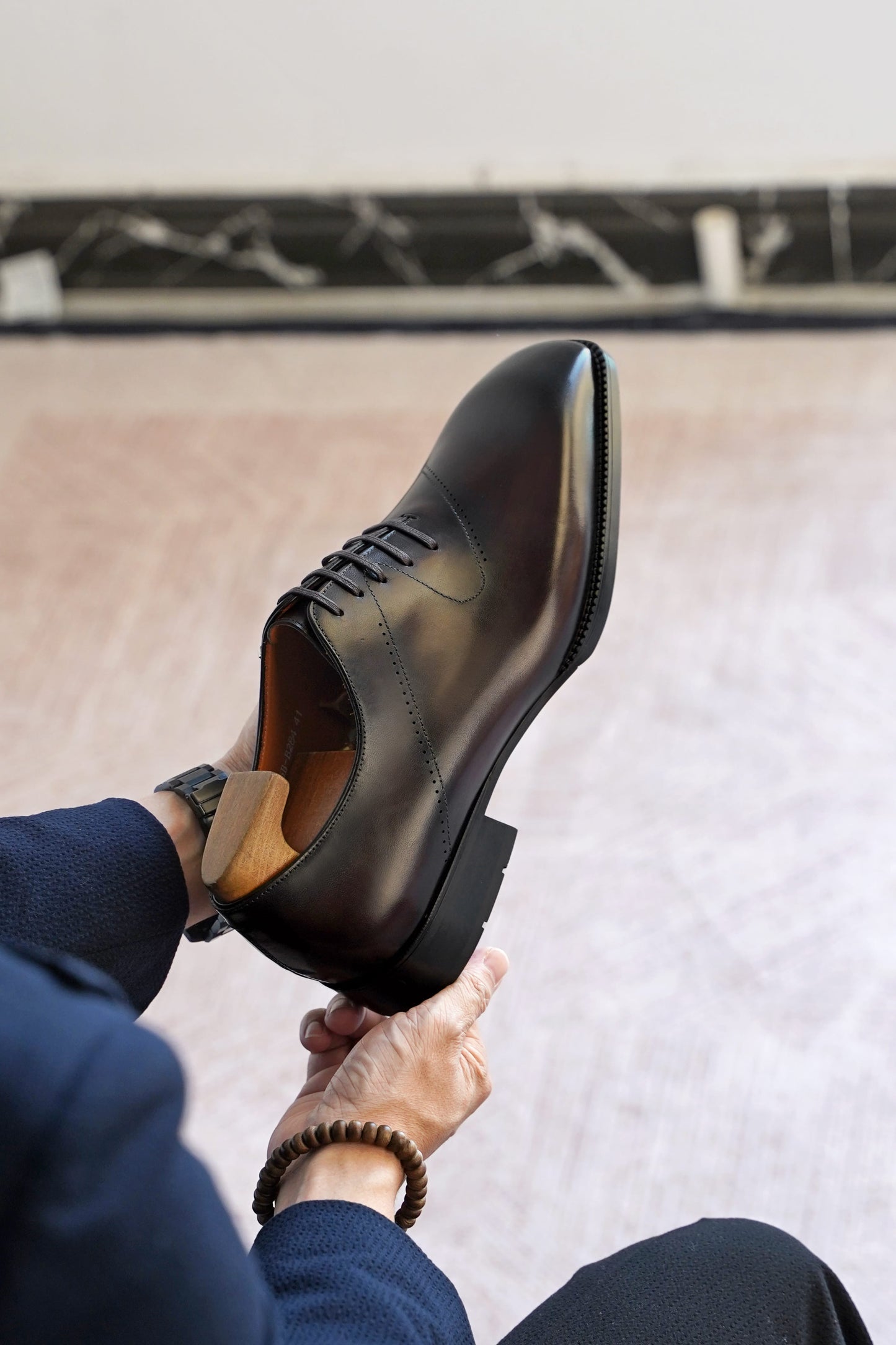 A close-up of a person holding up a polished dark brown leather lace-up dress shoe, showing the side profile and toe, with their hands and legs visible against a light indoor floor background.