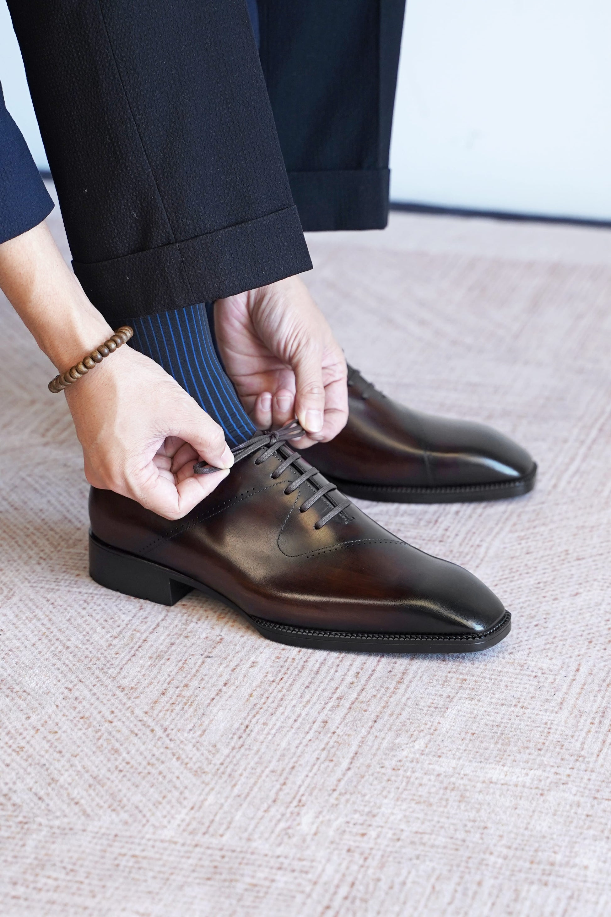 A close-up of a person's hands adjusting the laces of a dark brown oxford dress shoe, with black trousers and blue striped socks visible, set against a textured light beige carpet.