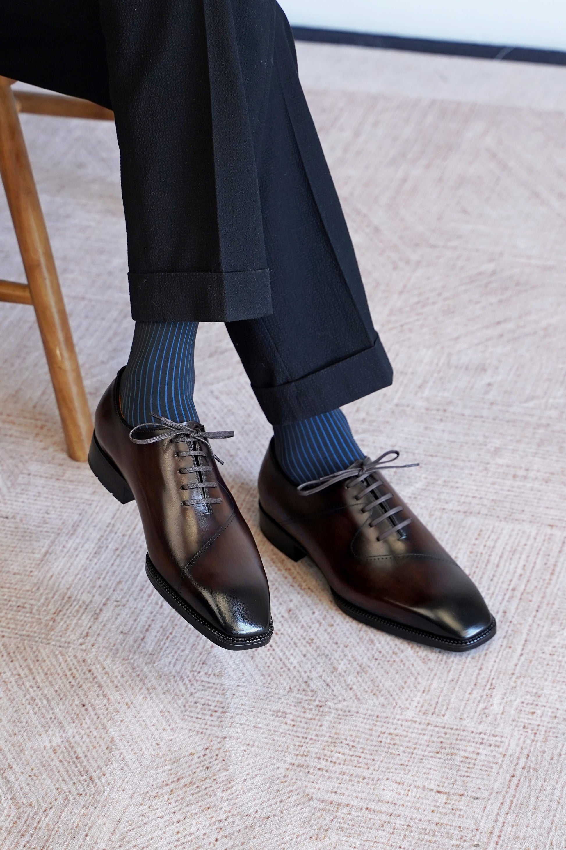 A close-up of a person's legs wearing dark brown oxford dress shoes with blue striped socks and black trousers, sitting on a light beige textured rug.