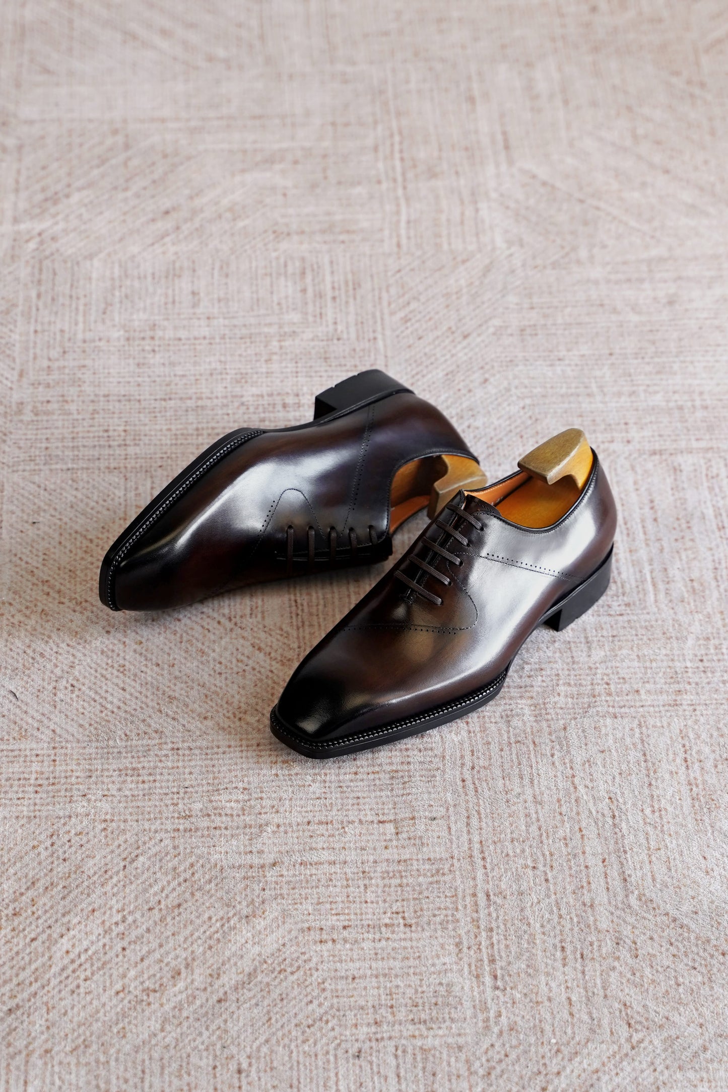 A pair of dark brown polished oxford dress shoes with brogue detailing, displayed on a textured beige woven rug.