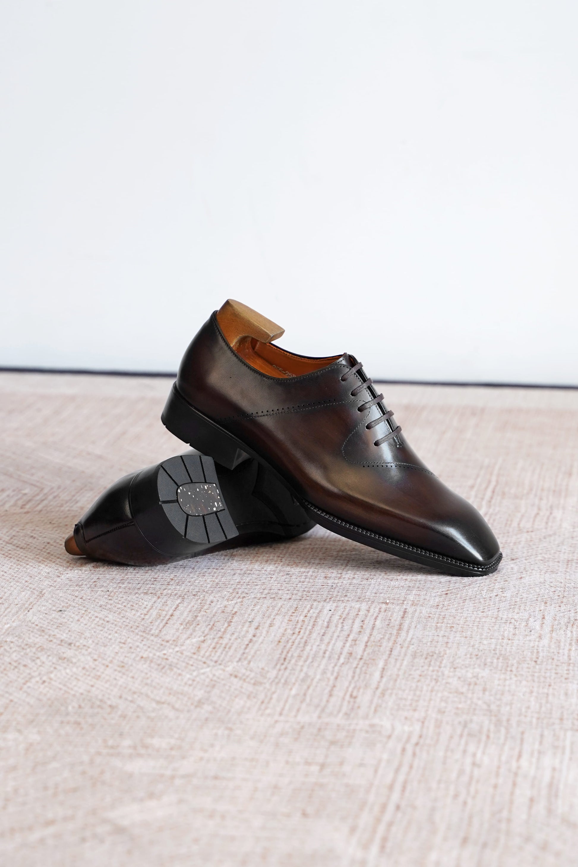 A pair of dark brown leather lace-up dress shoes with a glossy finish, displayed on a textured beige surface against a plain white background.