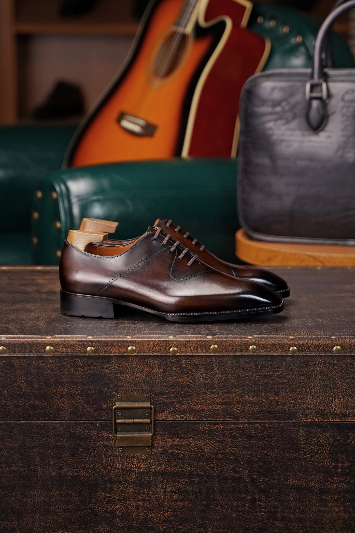 A pair of dark brown leather lace-up dress shoes with wooden shoe trees inside, displayed on a rustic wooden trunk with a guitar and a black leather handbag in the background.