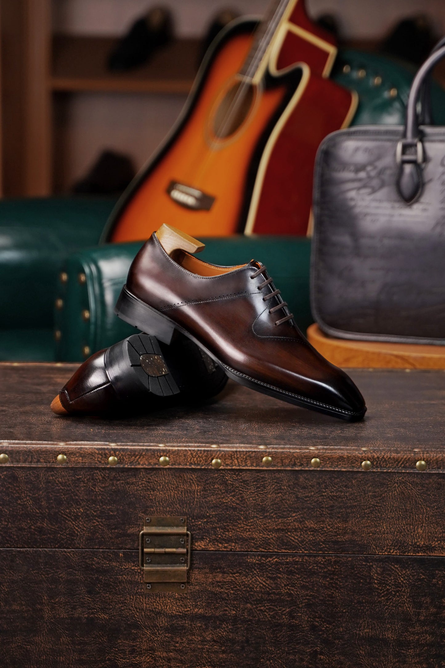 A pair of dark brown leather oxford dress shoes with black toe caps, displayed on a rustic wooden trunk with a guitar and a handbag in the background.