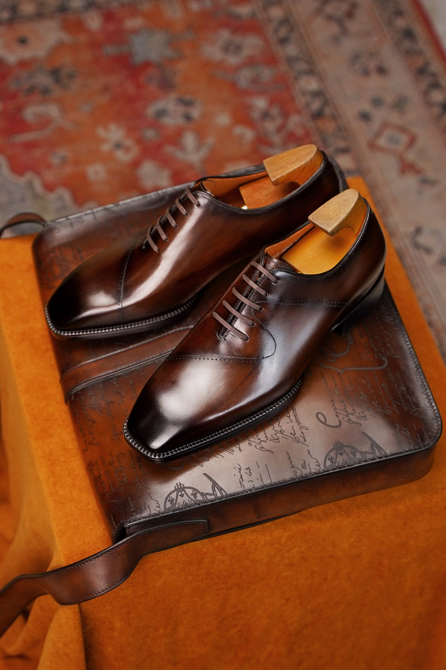 A pair of dark brown oxford dress shoes with wingtip caps and brogue detailing, displayed on a brown leather shoe box with a patterned rug in the background.