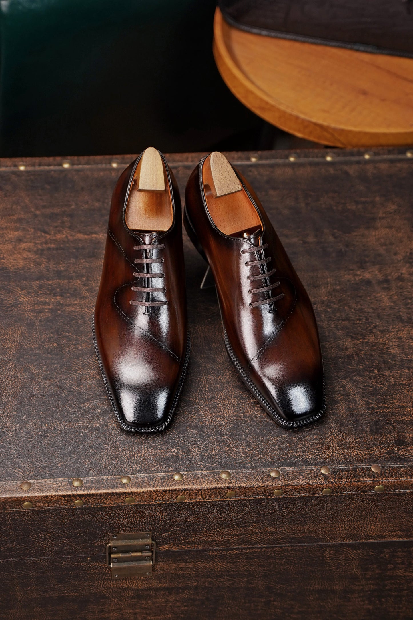 A pair of polished dark brown leather lace-up dress shoes with a glossy finish, displayed on a rustic dark wooden trunk.