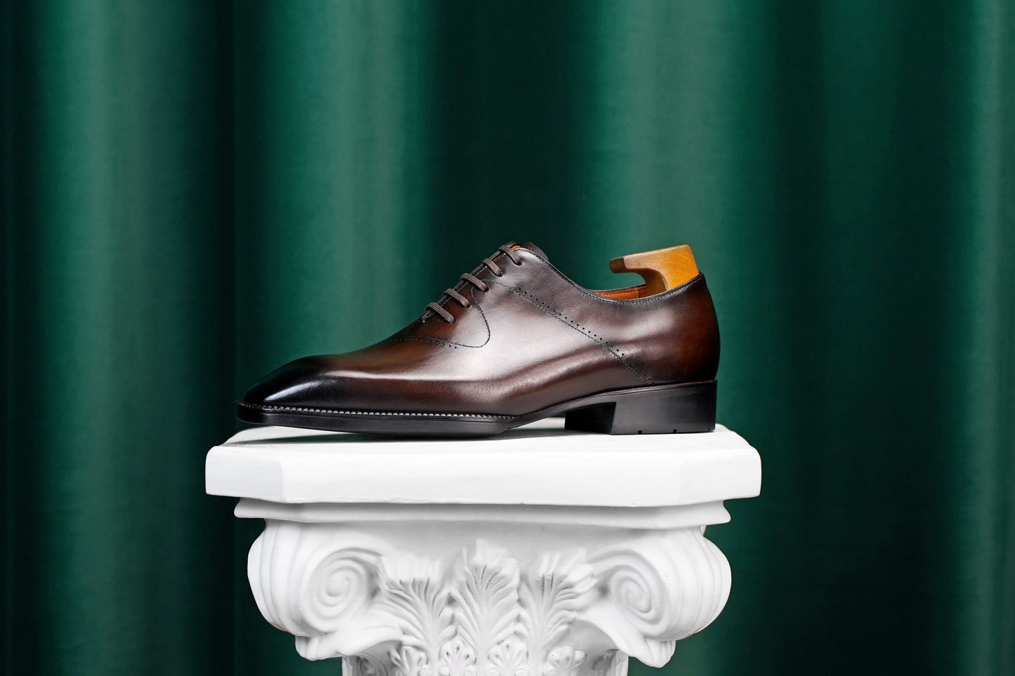A dark brown leather oxford dress shoe with a glossy toe and a light wood shoe tree inserted, displayed on a white decorative pedestal against a dark green curtain background.