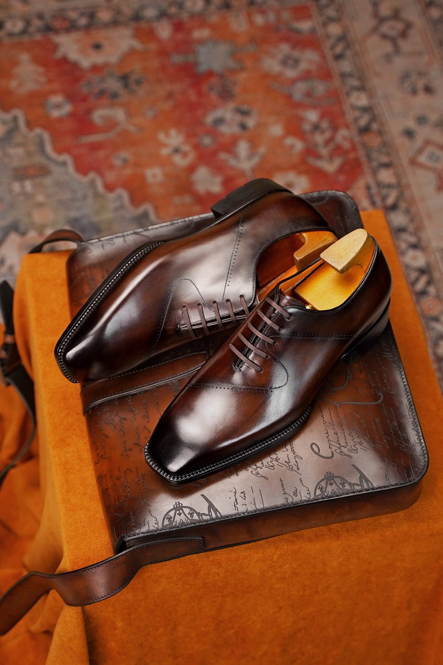 A pair of dark brown leather oxford dress shoes with a wingtip brogue design, displayed on a brown leather travel bag with a yellow shoe tree inside one shoe, set against an orange fabric surface and a patterned rug.